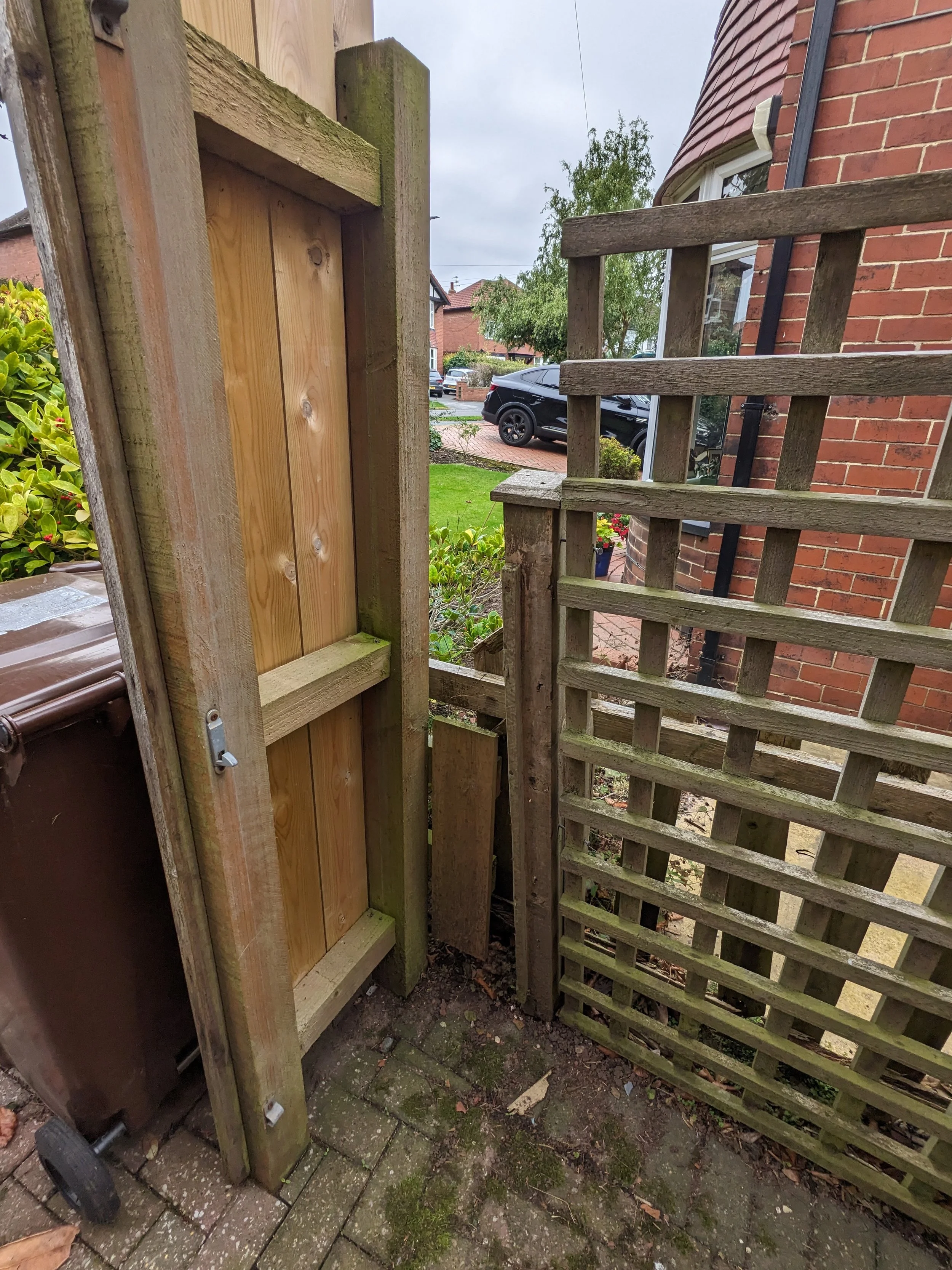 A small wooden garden gate with a latch, between a brick house and a wooden fence, leading into a yard with grass and trees.