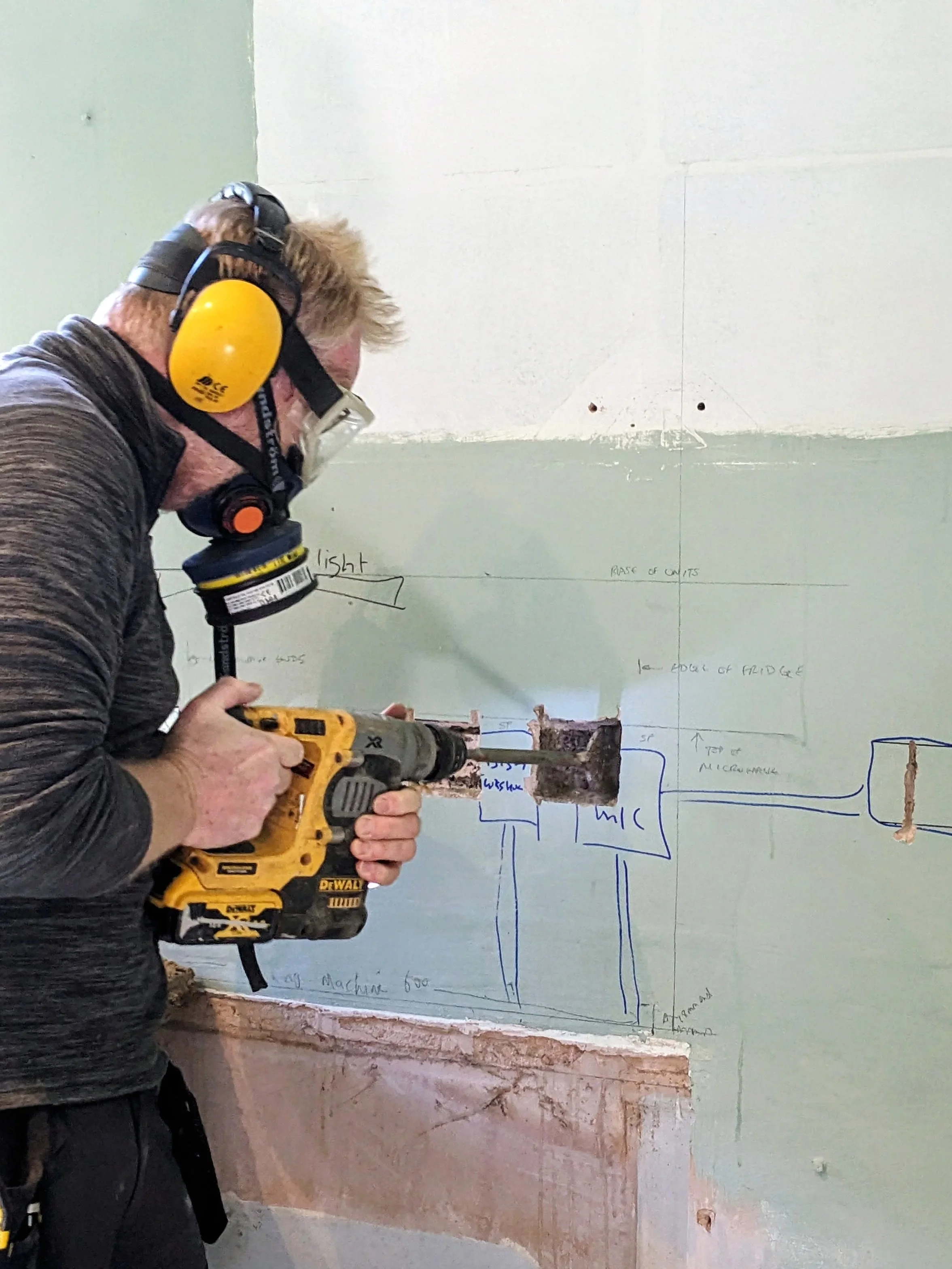 A man wearing protective gear, including yellow ear muffs, goggles, and a dust mask, using a cordless drill on a wall with handwritten blueprints or sketches, in a construction or renovation setting.