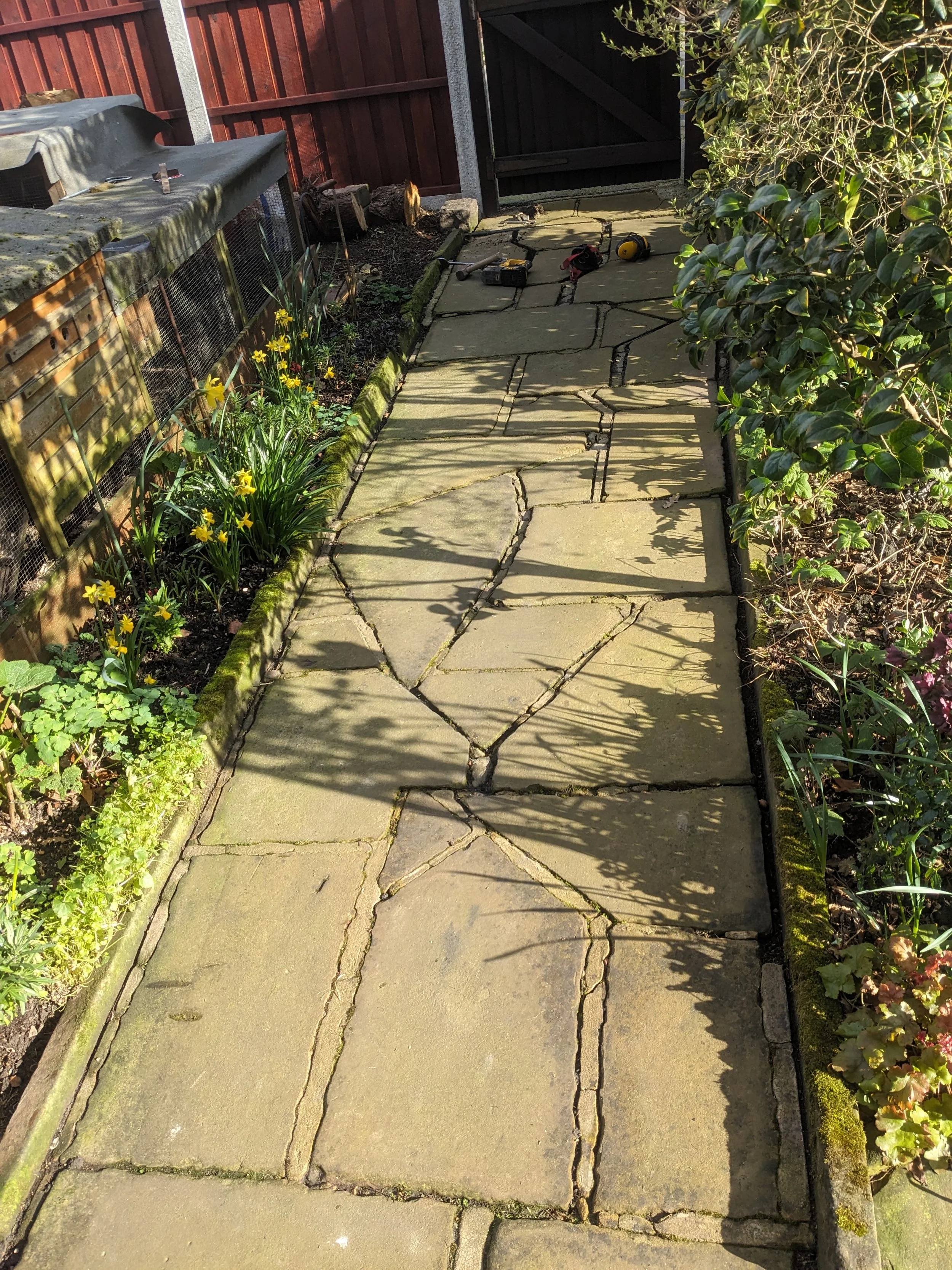 A stone pathway in a garden with flower beds on both sides, tools scattered at the far end, and a wooden fence with a gate in the background.