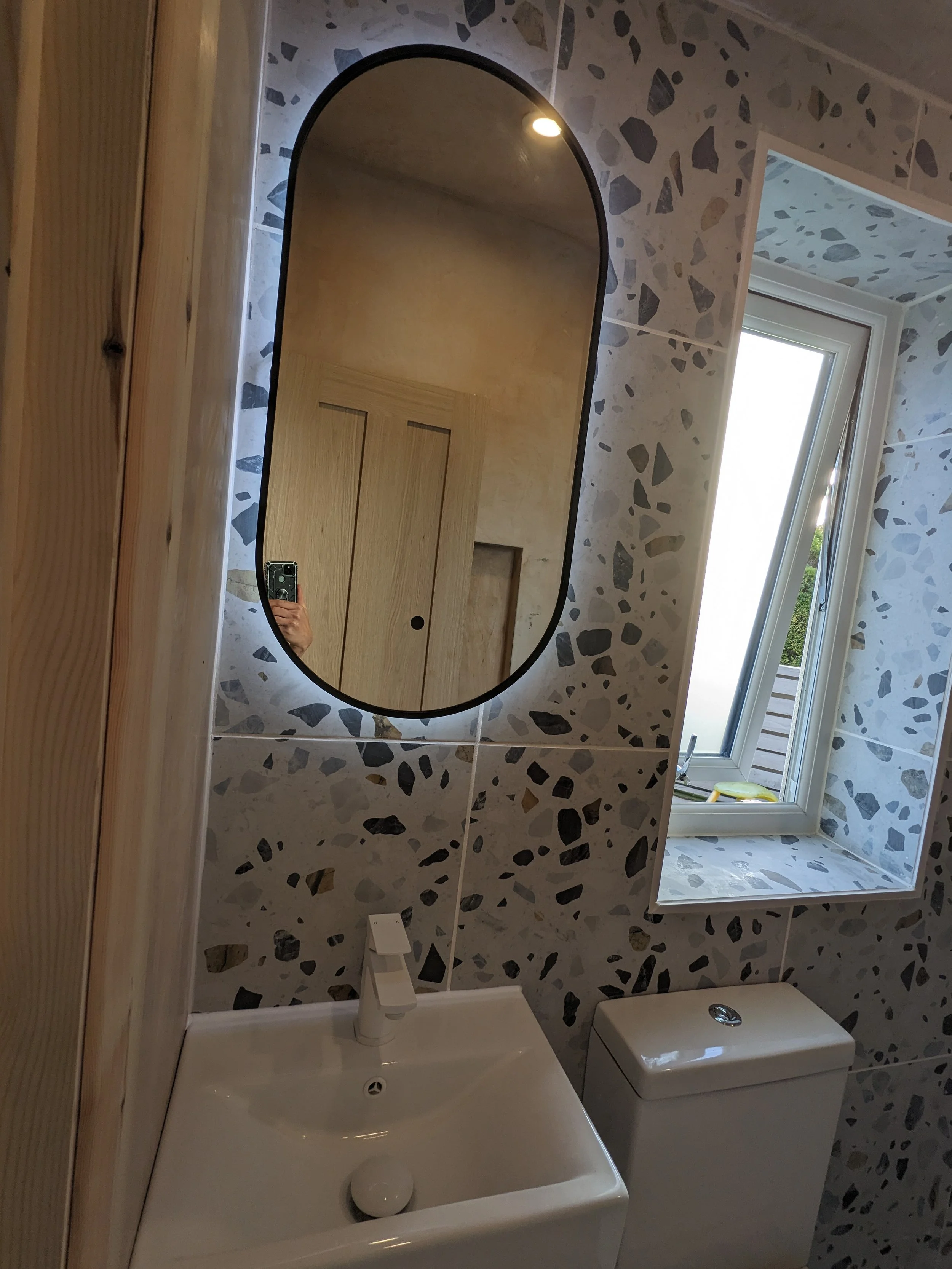 A bathroom with a terrazzo wall, a small white sink with a faucet, a white toilet, and a window with a white frame bringing in natural light.