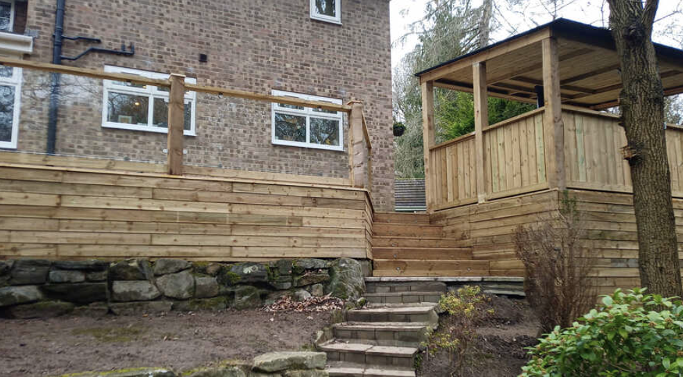 A newly constructed wooden deck with stairs and railing attaches to a brick house, with a small wooden gazebo nearby, surrounded by trees and bushes.