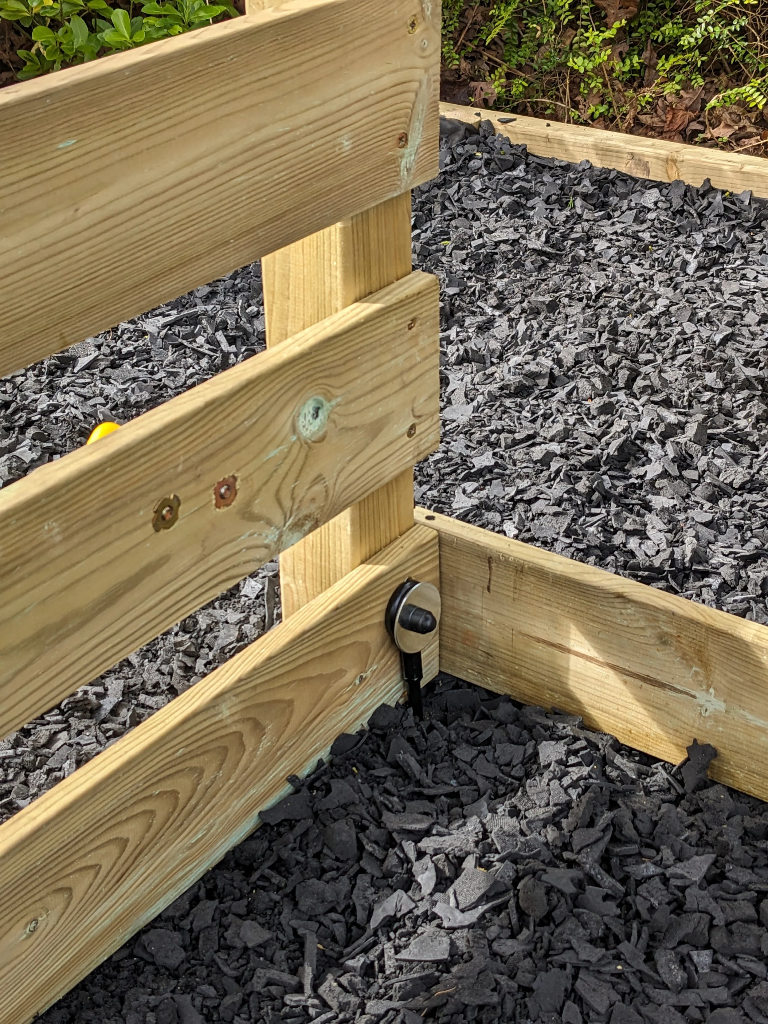 Close-up of a wooden garden bed frame with black landscape fabric or plastic edging beneath it, secured by a magnet and a black plastic clip.