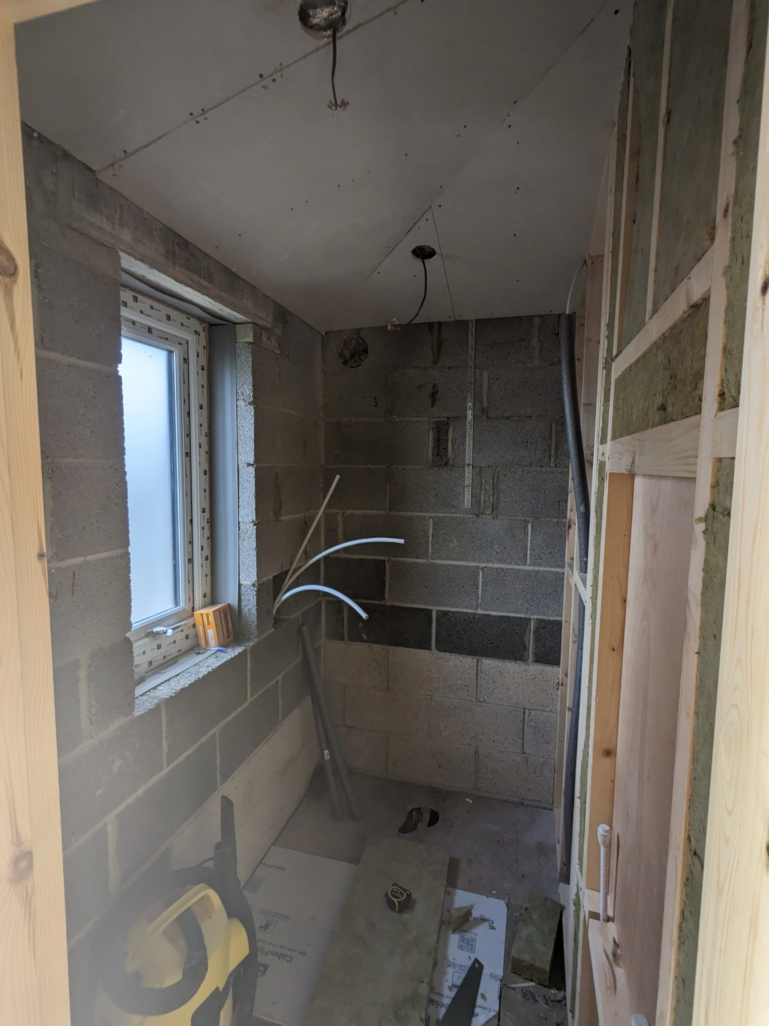 A small, unfinished bathroom or utility room under construction with concrete block walls, a window, exposed wiring, and construction tools and materials.