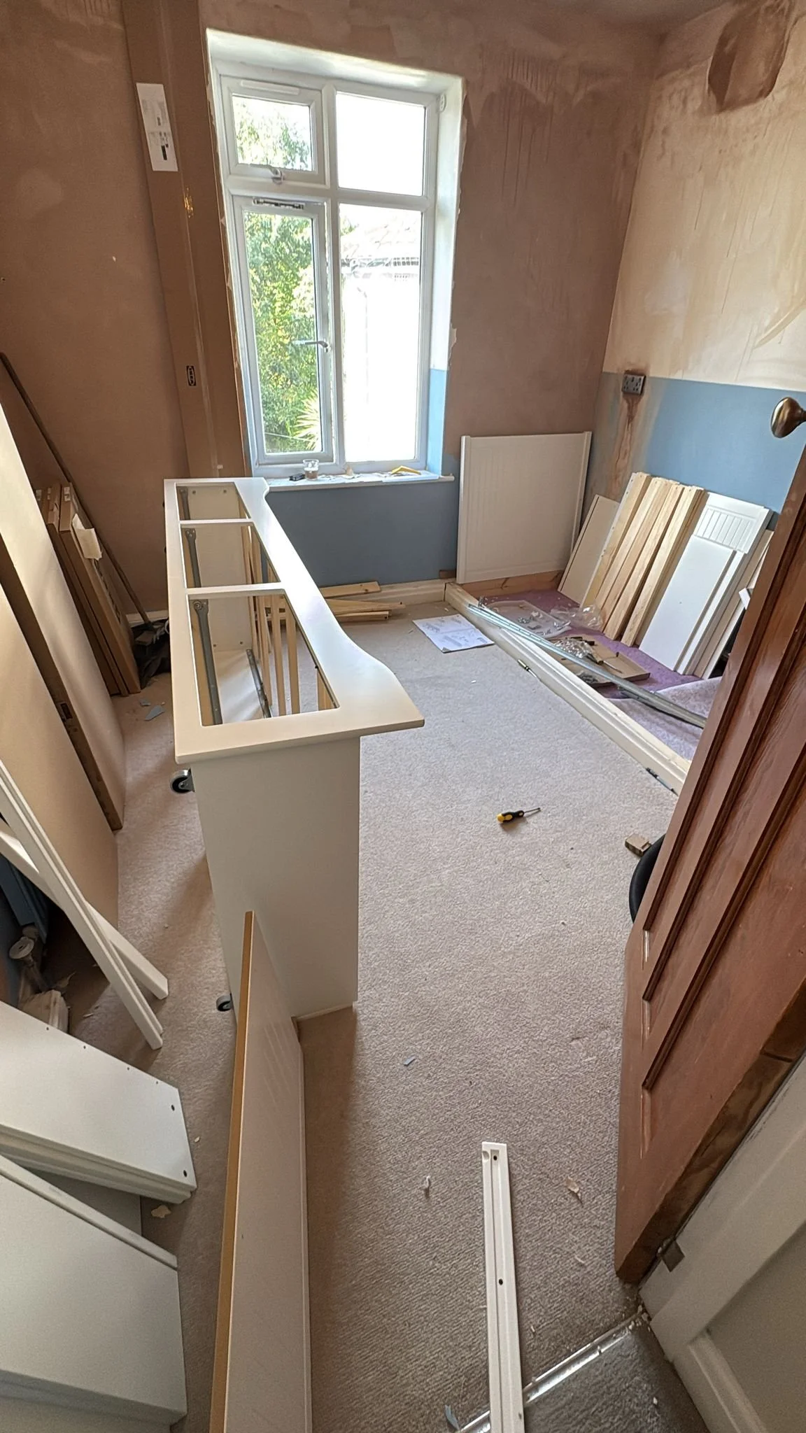 Room under renovation with construction materials, a partially assembled white cabinet, and scattered tools, with a window showing greenery outside.