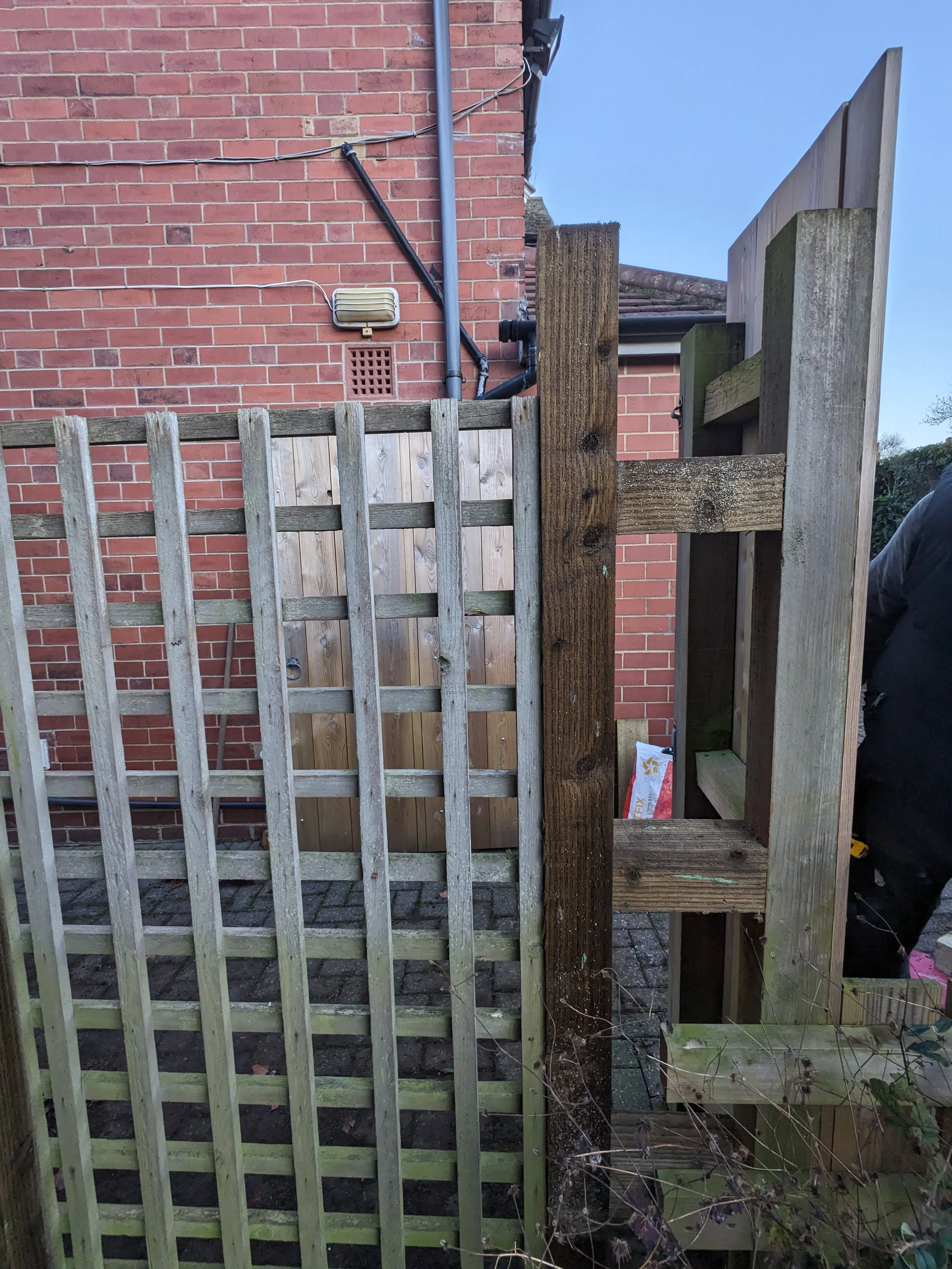 A partially constructed wooden fence with a brick building wall in the background. The fence features vertical and horizontal wooden planks, with some sections unfinished and wooden beams visible. There are outdoor utility fixtures on the brick wall,