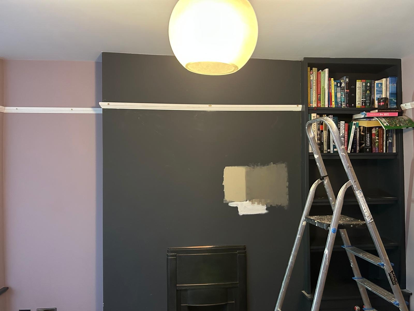 Interior of a room with a dark black wall, partially painted over, with a white chalk line above. There is a bookshelf filled with books and a step ladder in front of it, with a ceiling light overhead.