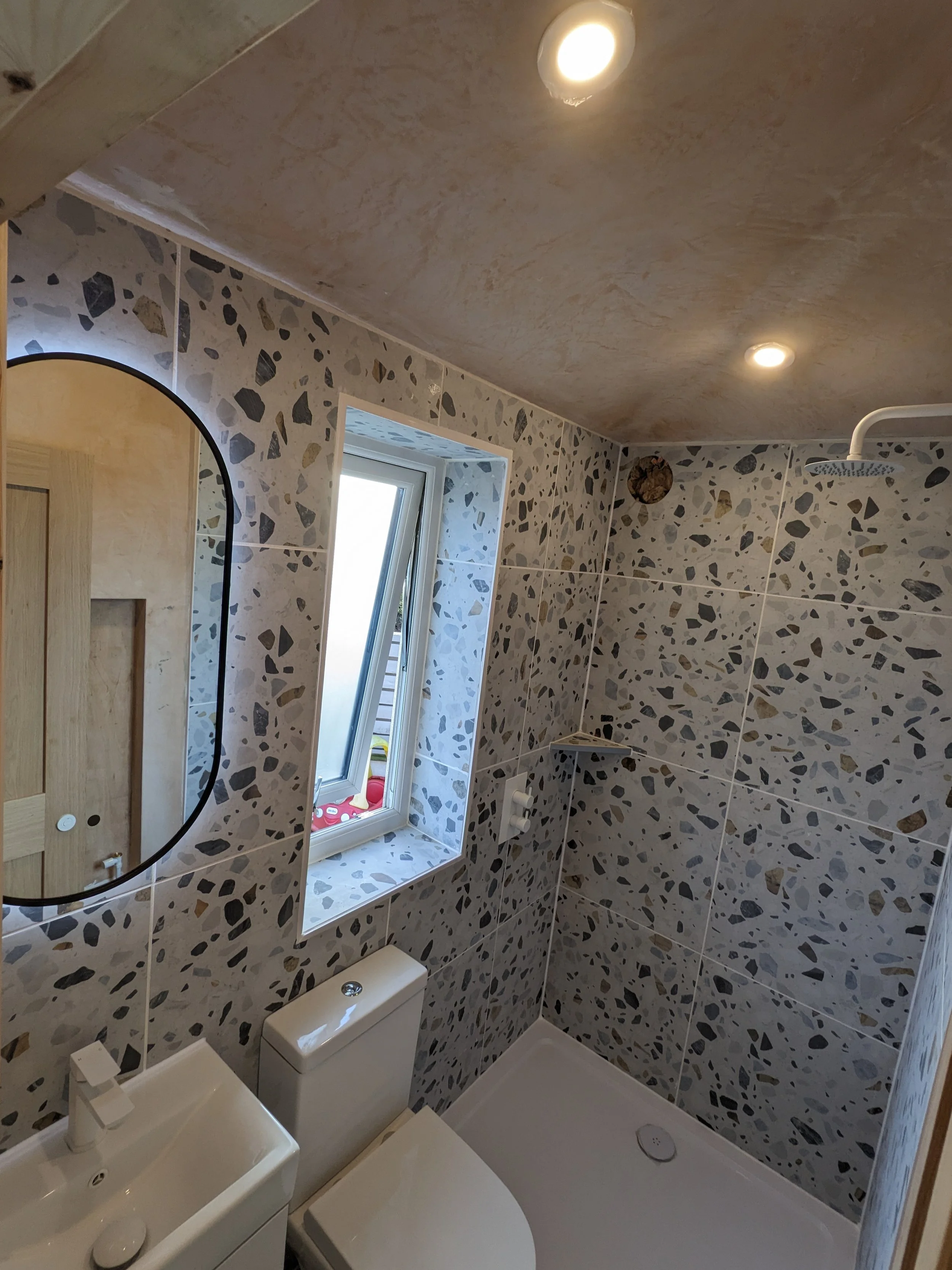 Newly installed Bathroom with terrazzo tile walls, a small white sink, a toilet, a window, and a shower area with a rain shower head. Recessed ceiling lights illuminate the room.