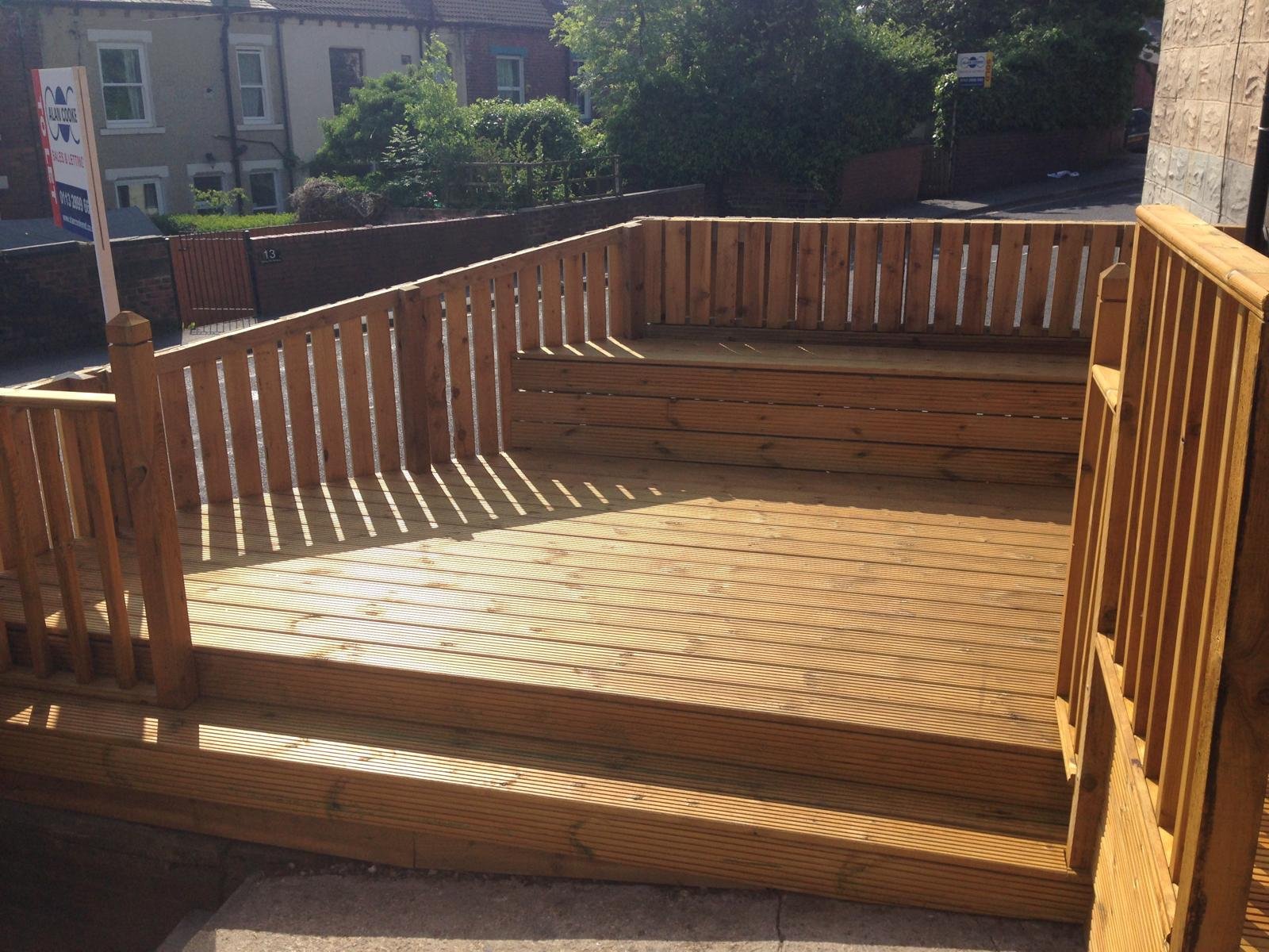 Empty wooden deck with a railing, located outdoors on a sunny day with houses and trees in the background.
