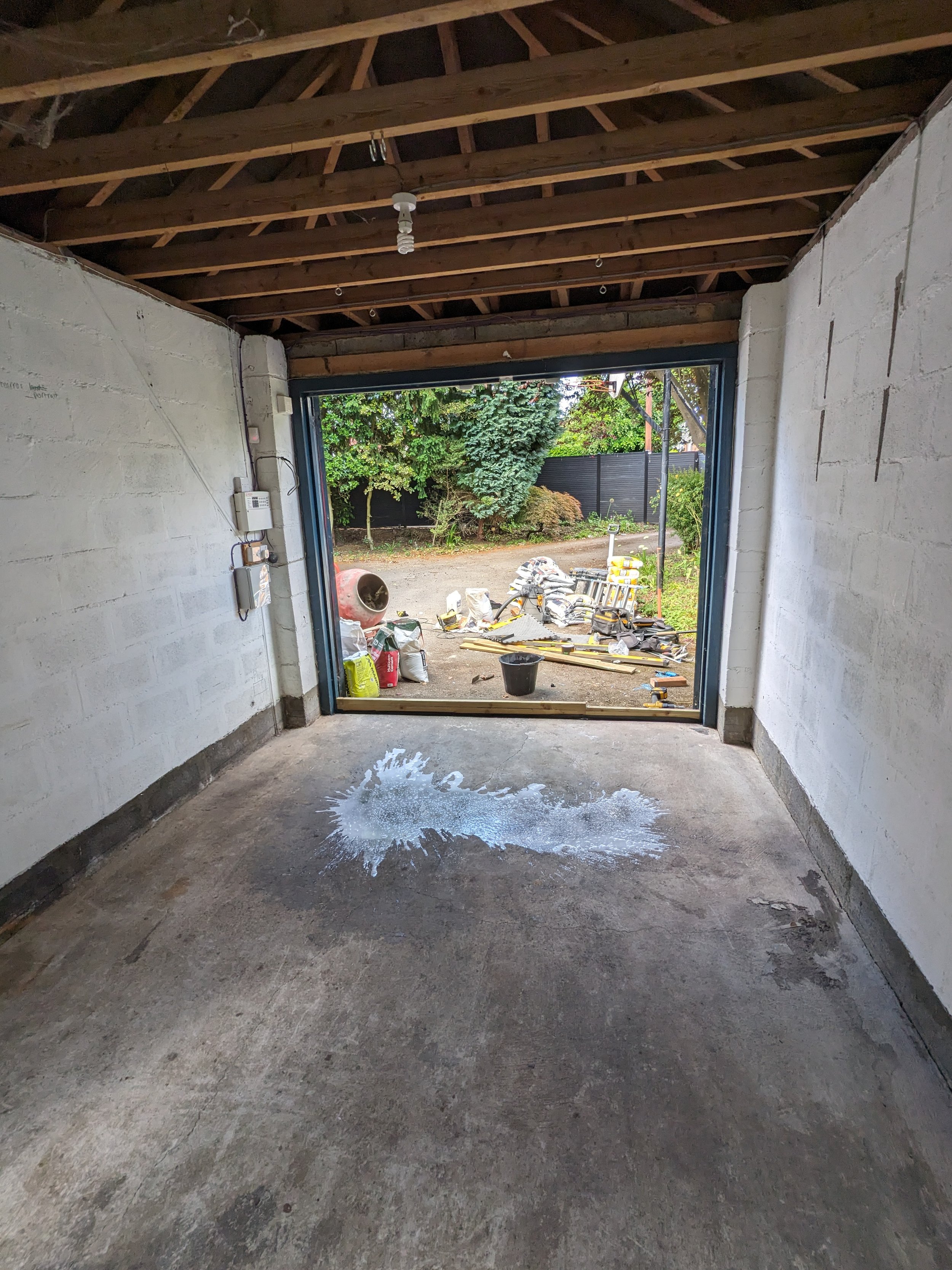 Inside a garage with an unfinished ceiling, concrete floor, and a pile of construction materials outside visible through an open garage door. There is a white paint spill on the floor.