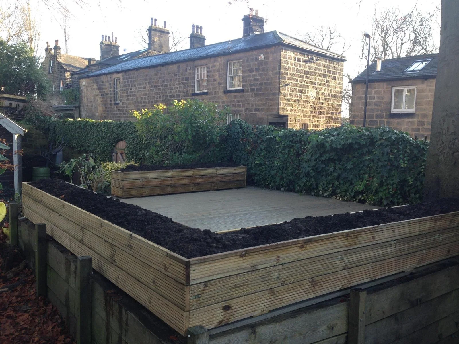 A backyard garden area with a wooden deck and empty flower beds bordered by wooden planks, with a large bush and neighboring stone houses in the background.