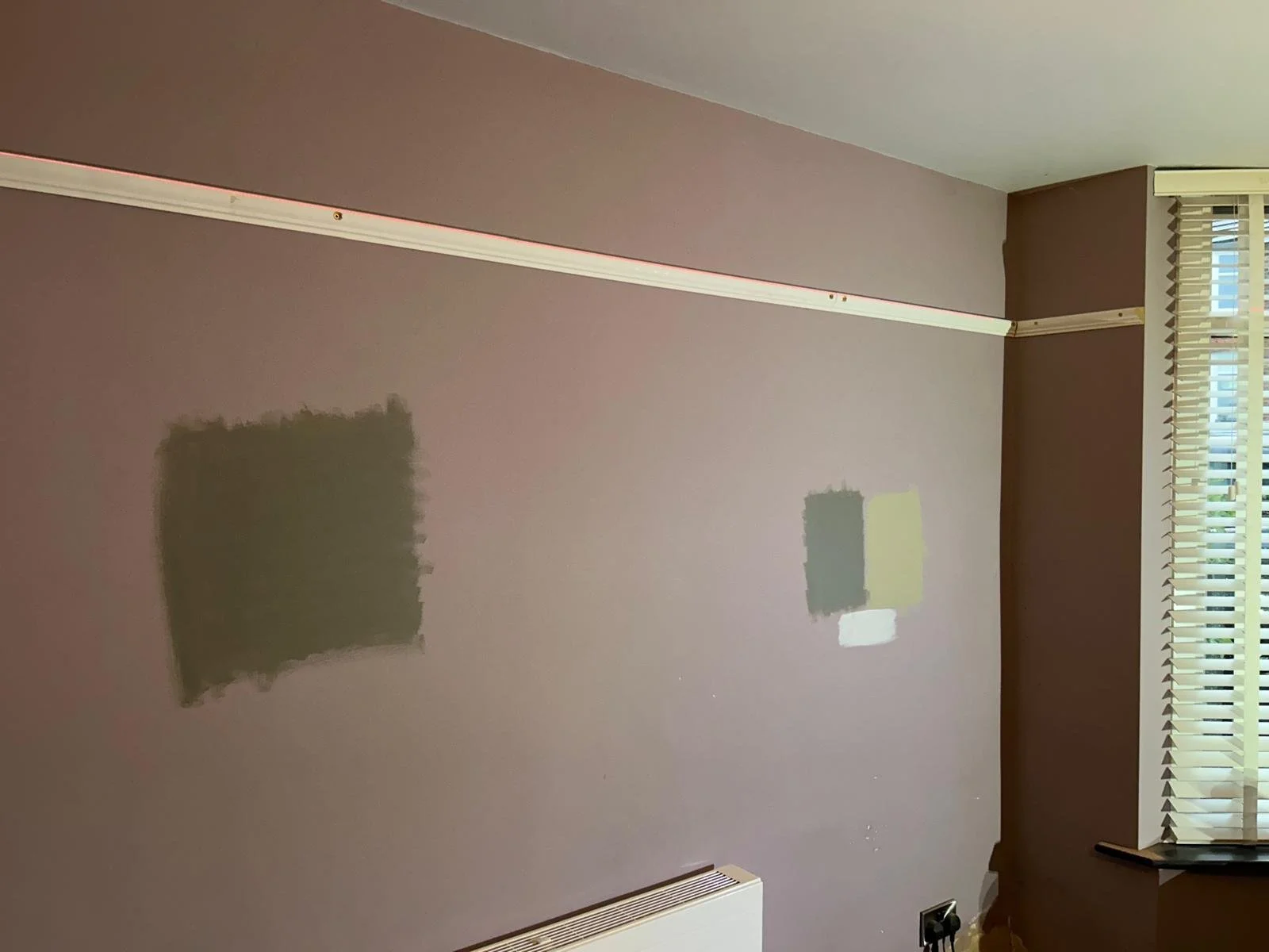 A wall with two painted patches, a window with blinds, and a heating unit at the bottom.