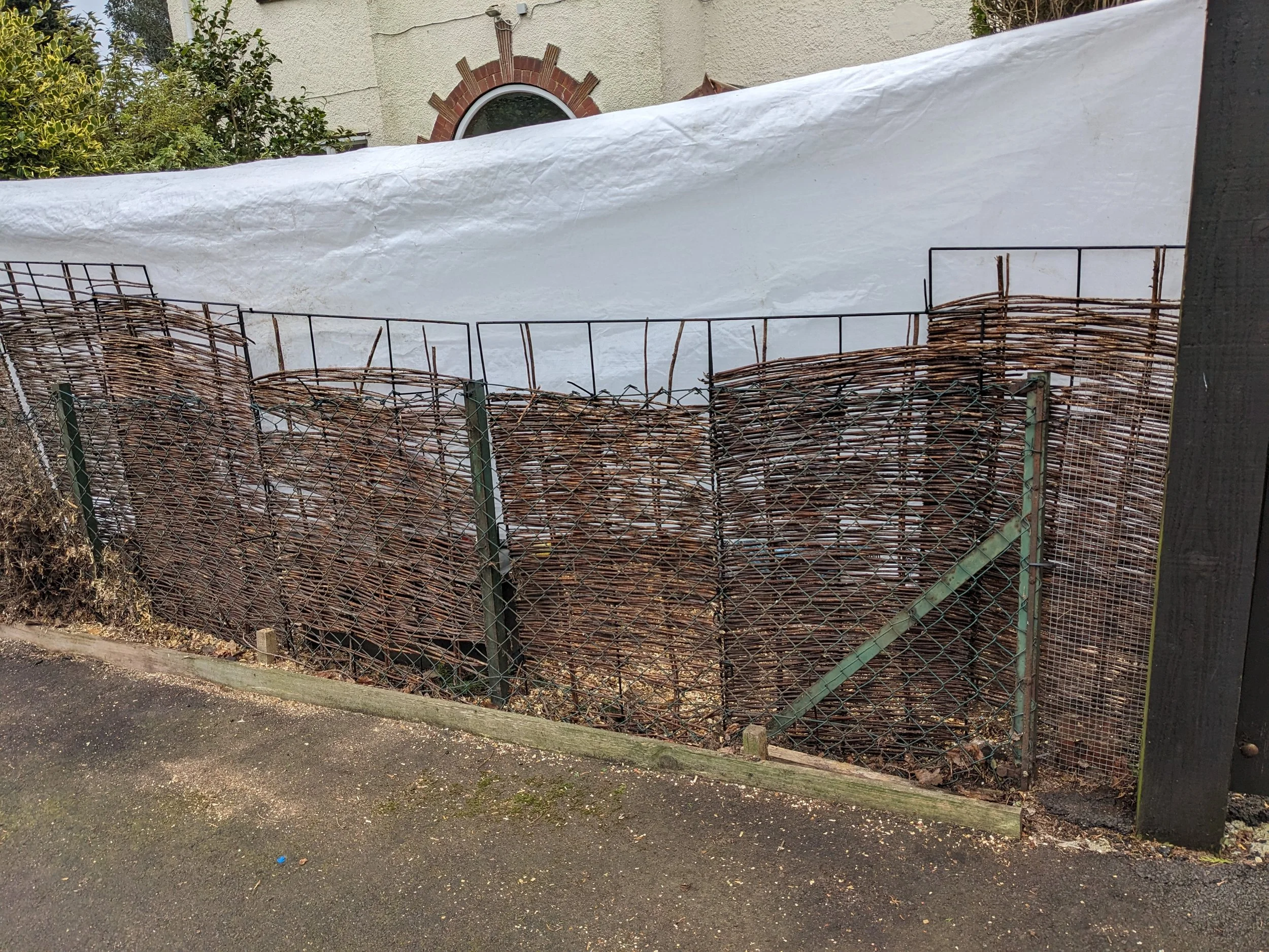 A metal wire fence with a lattice design and angled support in front of a pile of small logs or sticks behind a white tarp. Part of a building with a window and brick arch is visible in the background.