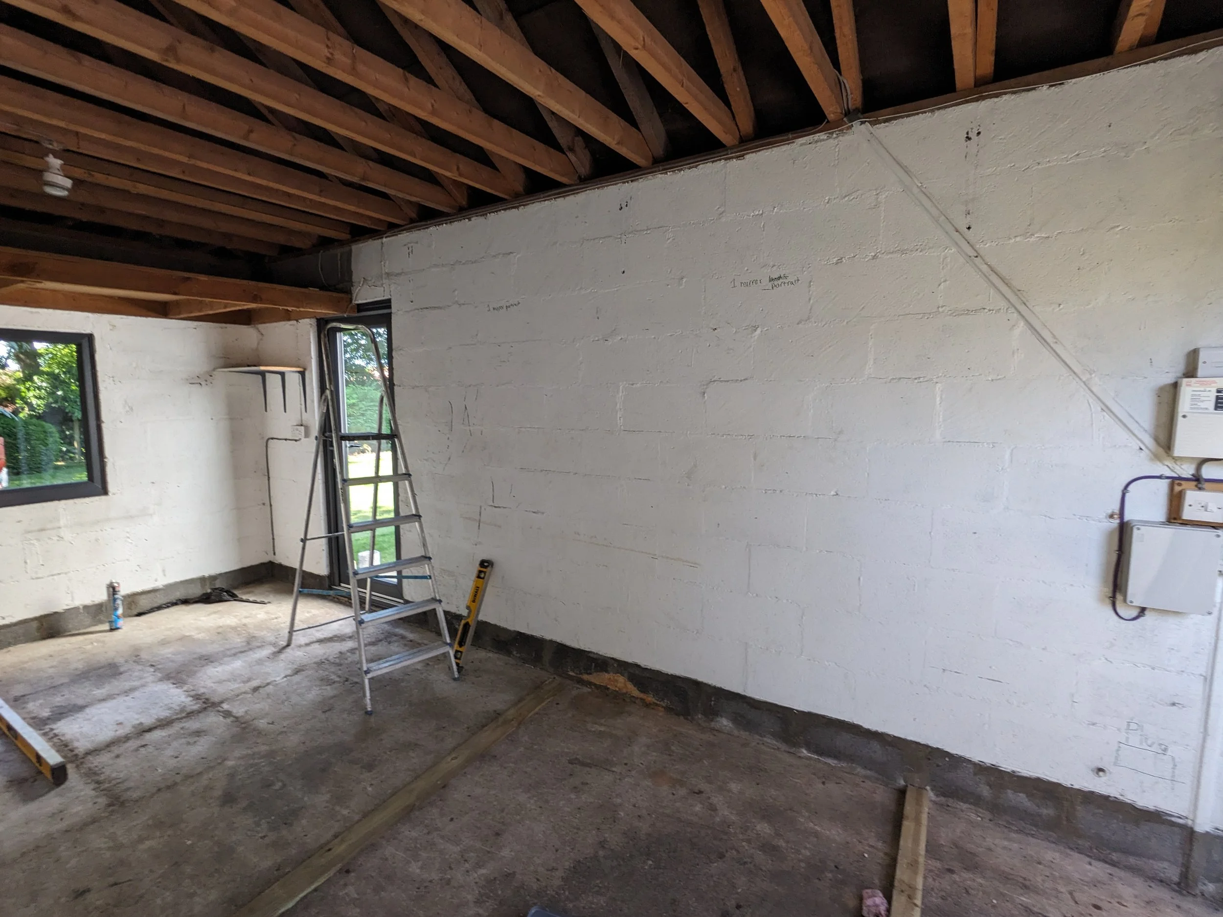 Interior of a room under construction with exposed ceiling beams, a white brick wall, an open door, and a ladder.