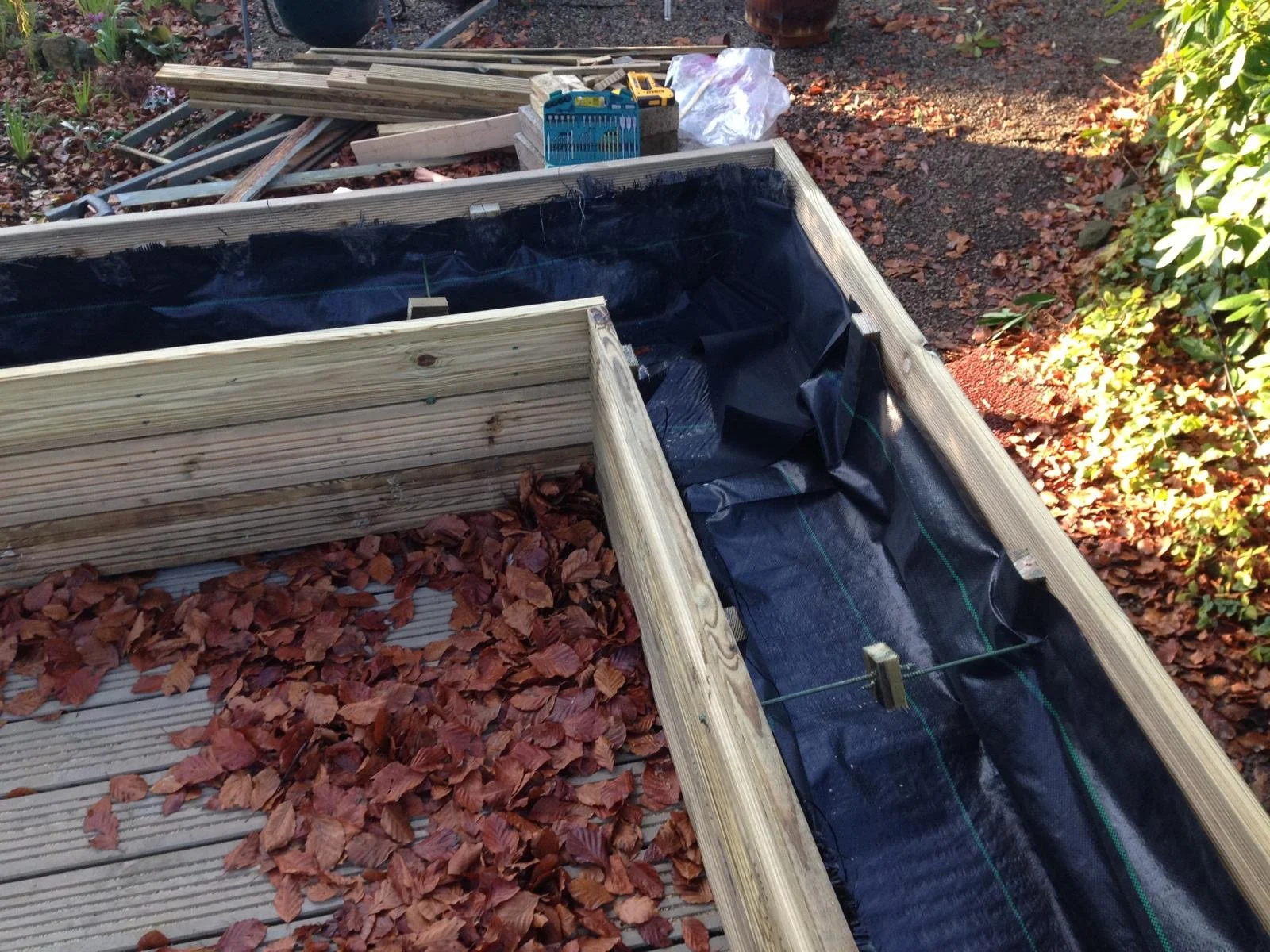 Wooden garden bed frame with black plastic liner, outdoors on a wooden deck, surrounded by autumn leaves and various gardening tools in the background.