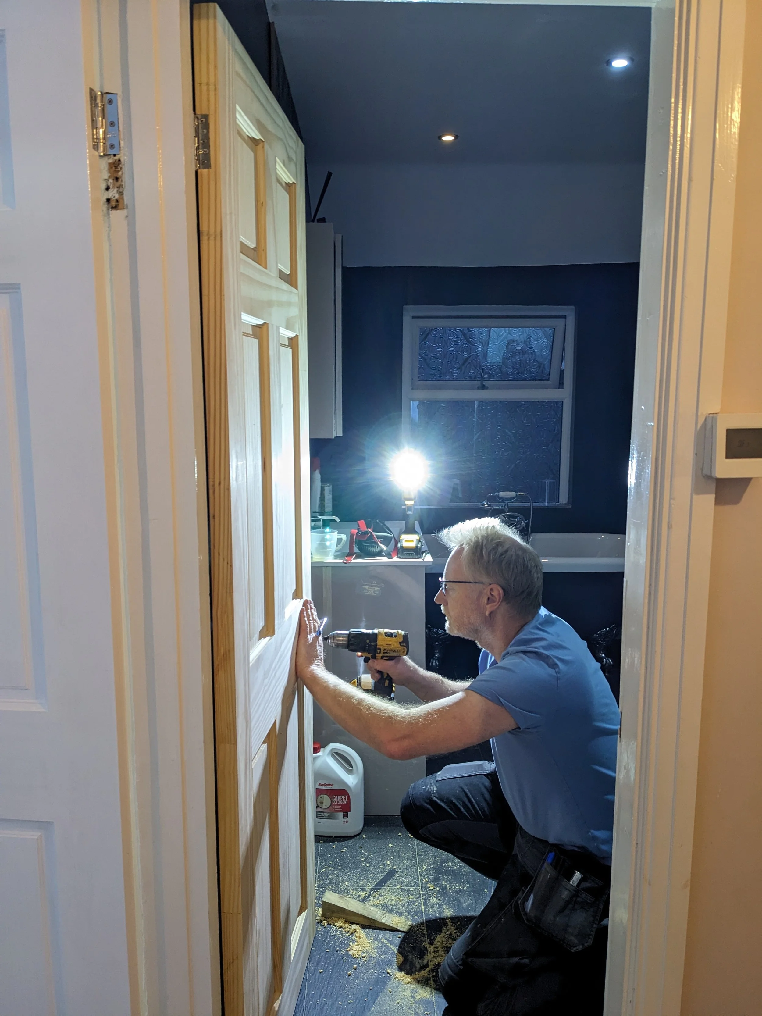 A man working on installing or repairing a wooden door in a room, using a power drill. The room appears to be a laundry or utility room with a sink, cleaning supplies, and containers visible in the background. The man is crouched down, wearing glasse