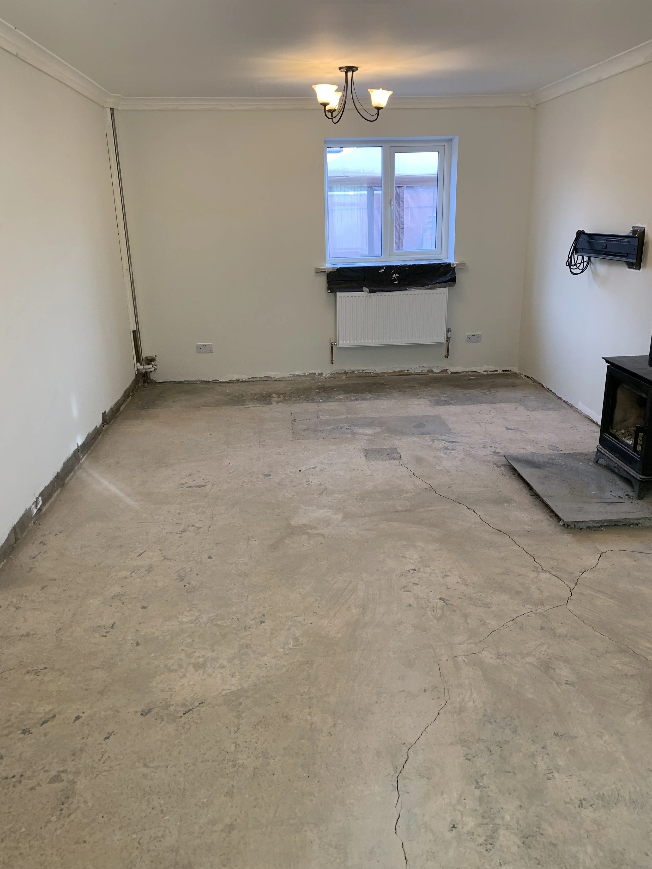 Room with unfinished floor, white walls, a ceiling light fixture, a window with black plastic covering, radiator, fireplace, and some exposed wiring.