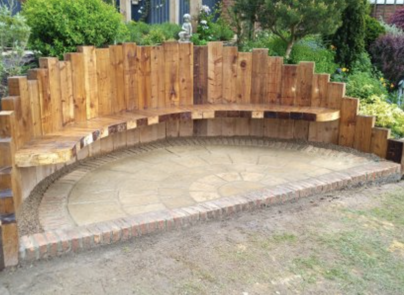 Curved outdoor wooden bench with backrest on a stone-paved patio, surrounded by lush green trees and bushes.