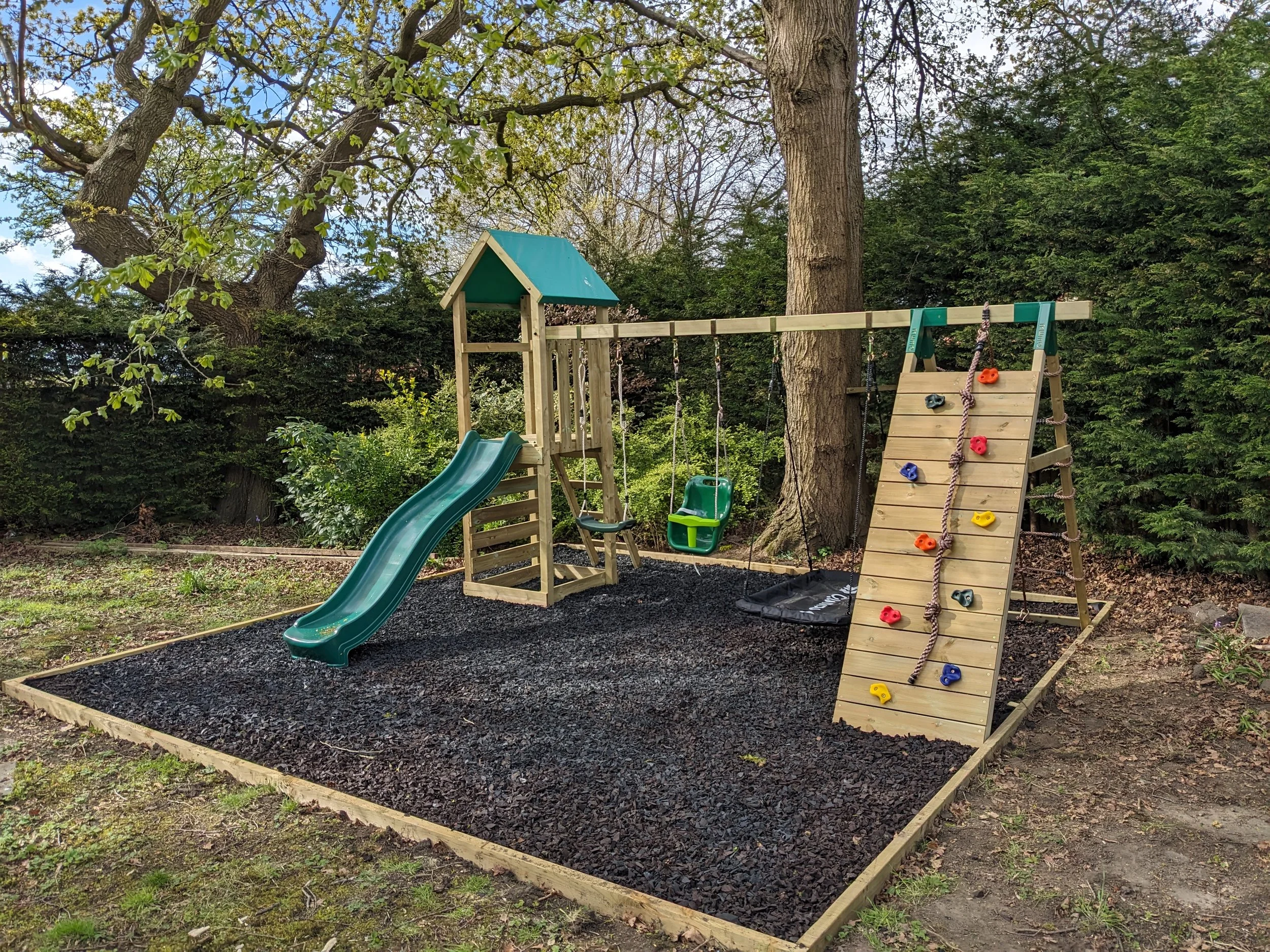Children's outdoor playset with slide, swing, climbing wall, and sandbox in a backyard surrounded by trees and greenery.