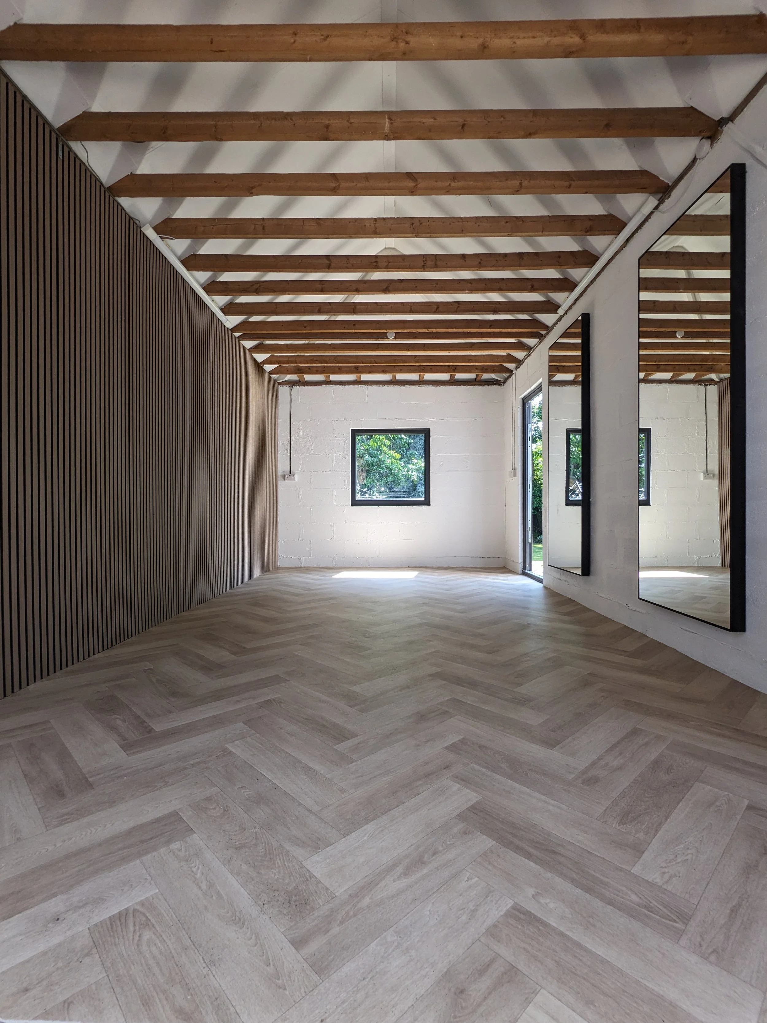 Refurbished garage gym room with white brick walls, wooden ceiling beams, large mirrors on the right, a window on the back wall, and wood-patterned flooring.