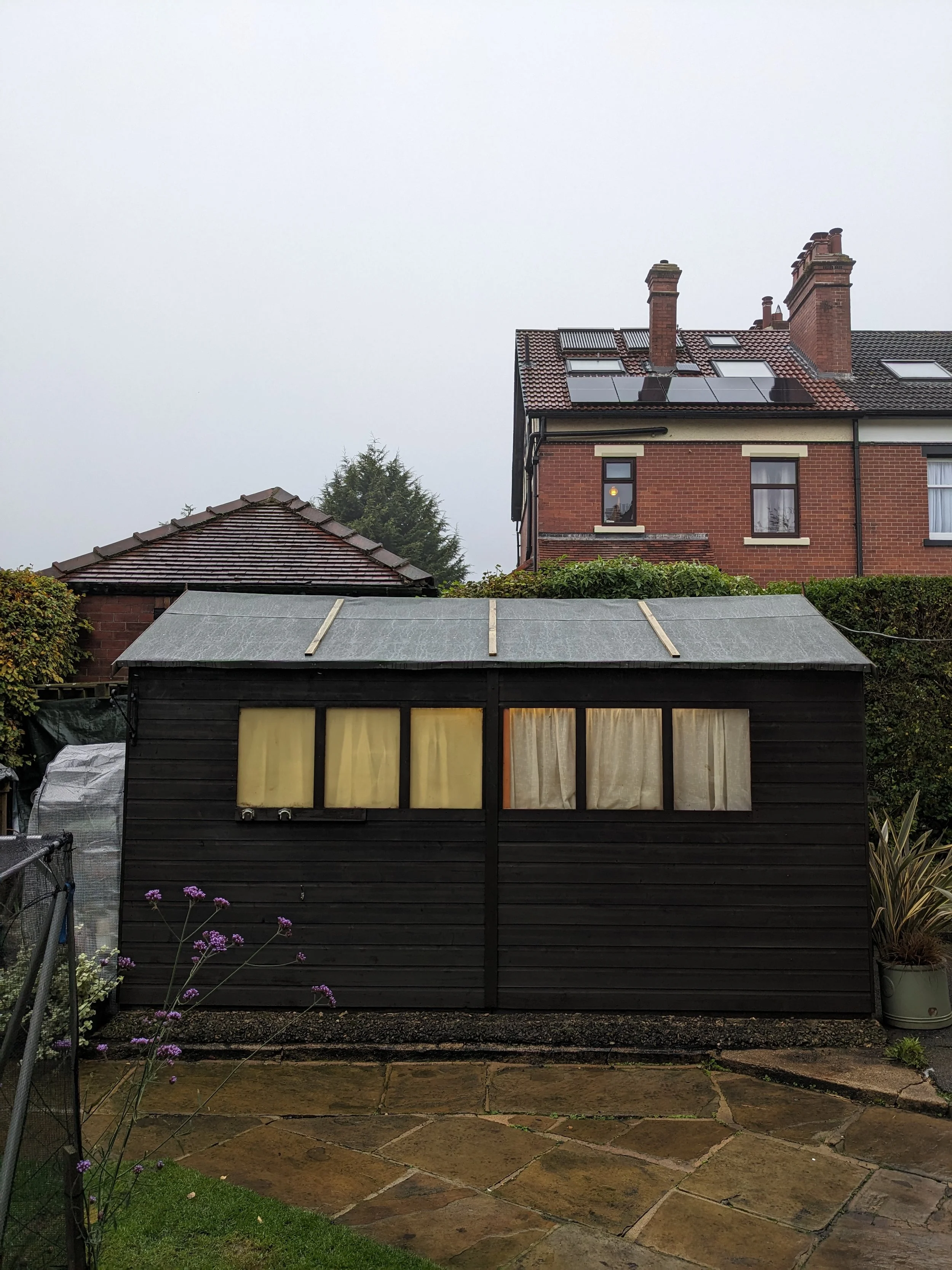 A backyard garden with a black wooden shed with yellow curtains, a stone patio, and a chained fence with flower plants in a pot and purple flowers in the foreground. A two-story brick house with solar panels on the roof is in the background under a c