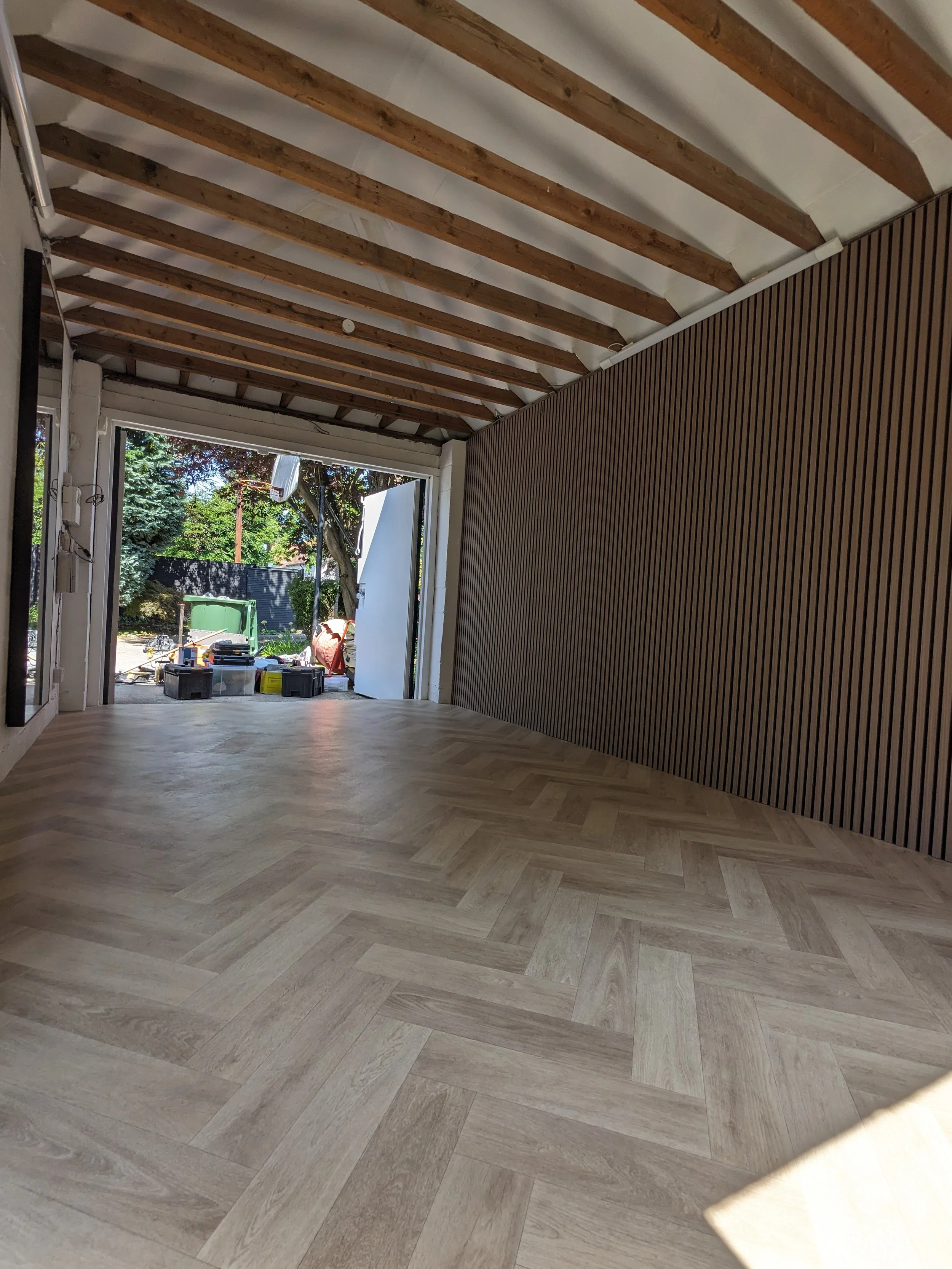 Interior view of a room with unfinished ceiling showing wooden beams, a wood-paneled wall, and light wood flooring, with an open garage door leading outside to trees and construction tools.
