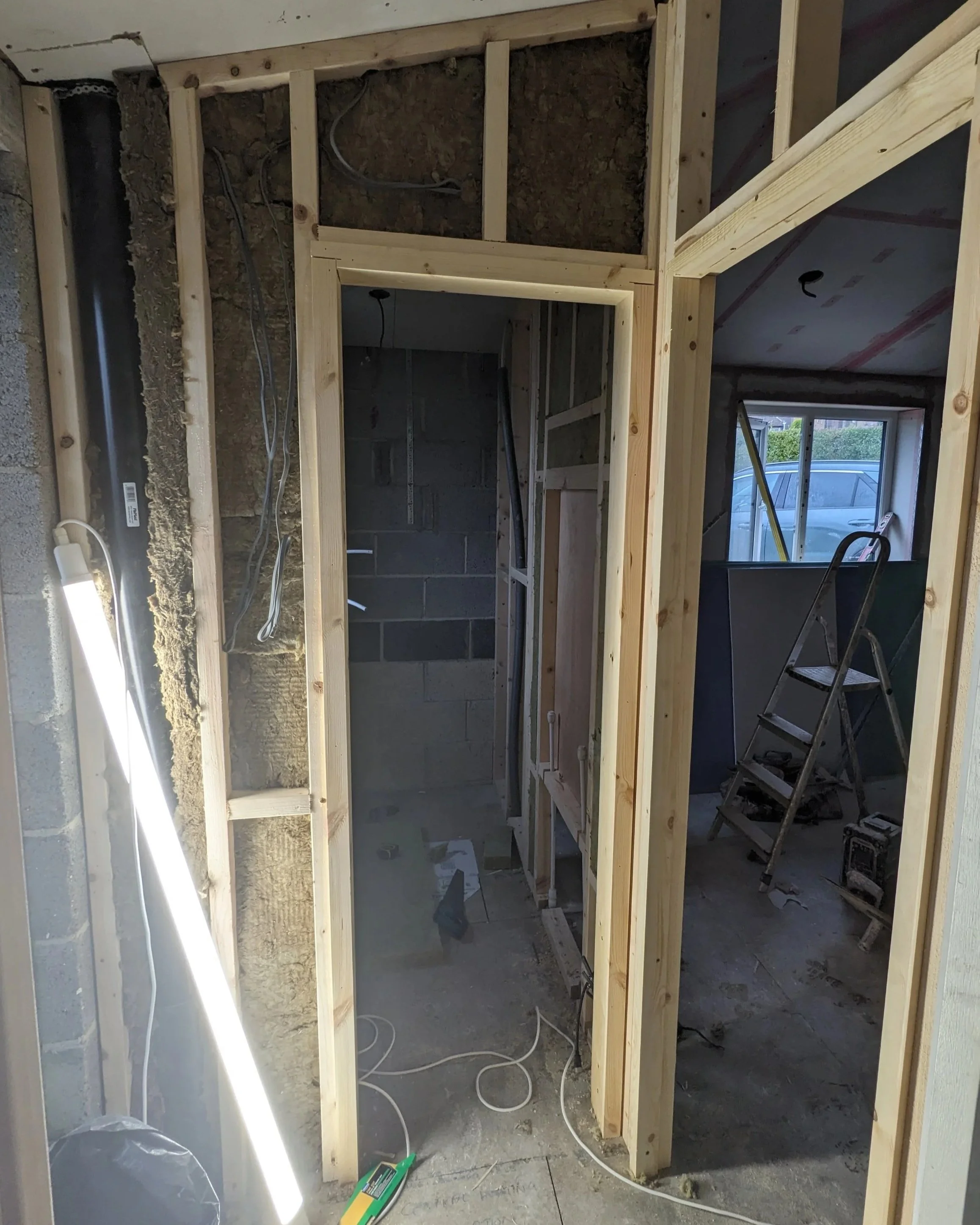 Interior view of a room under construction with exposed insulation, wooden framing, a step ladder, construction tools, and a window in the background.