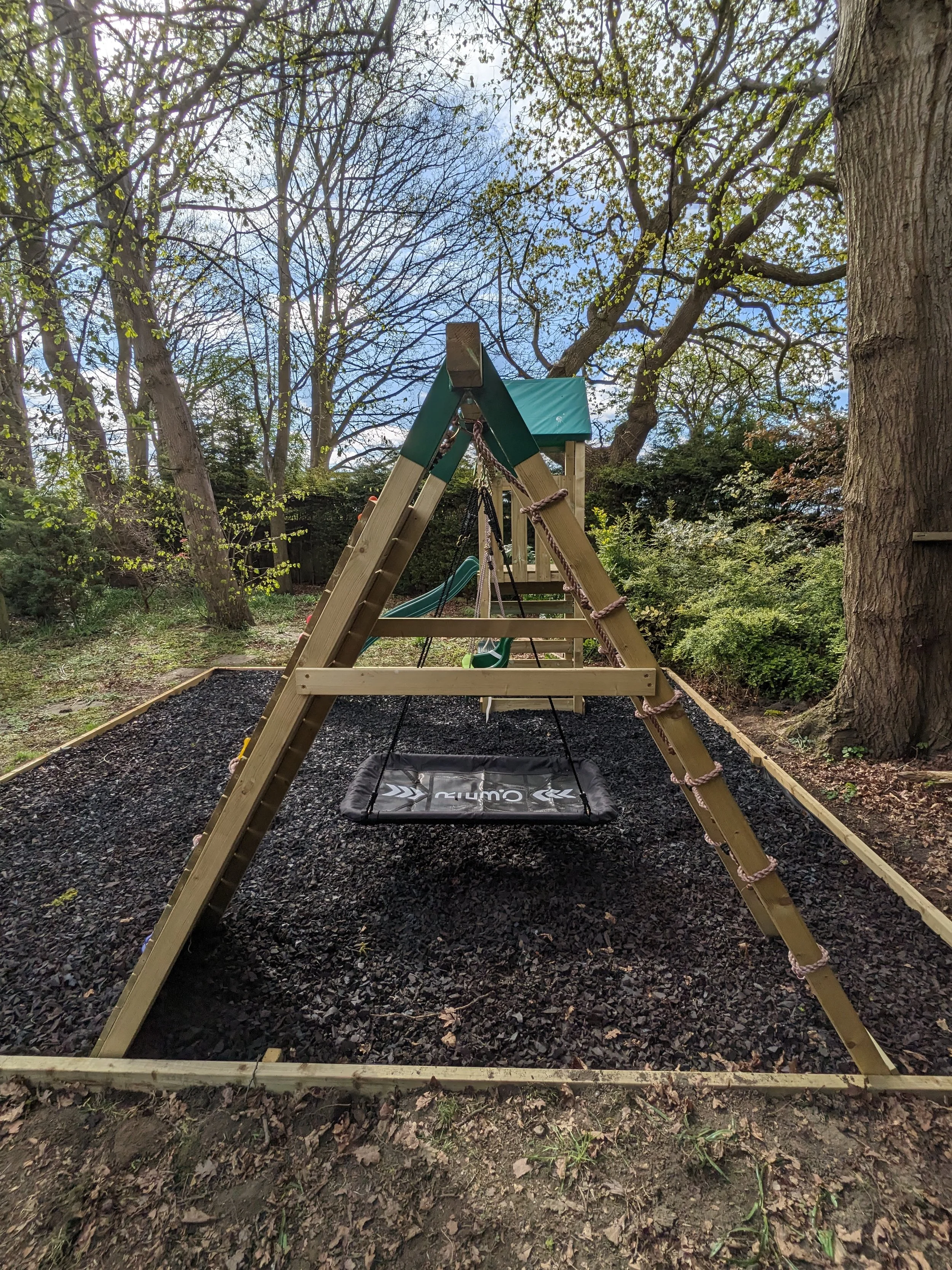 A children's backyard playset with a ladder, slide, and swing, set on a black mulch surface surrounded by trees and greenery.