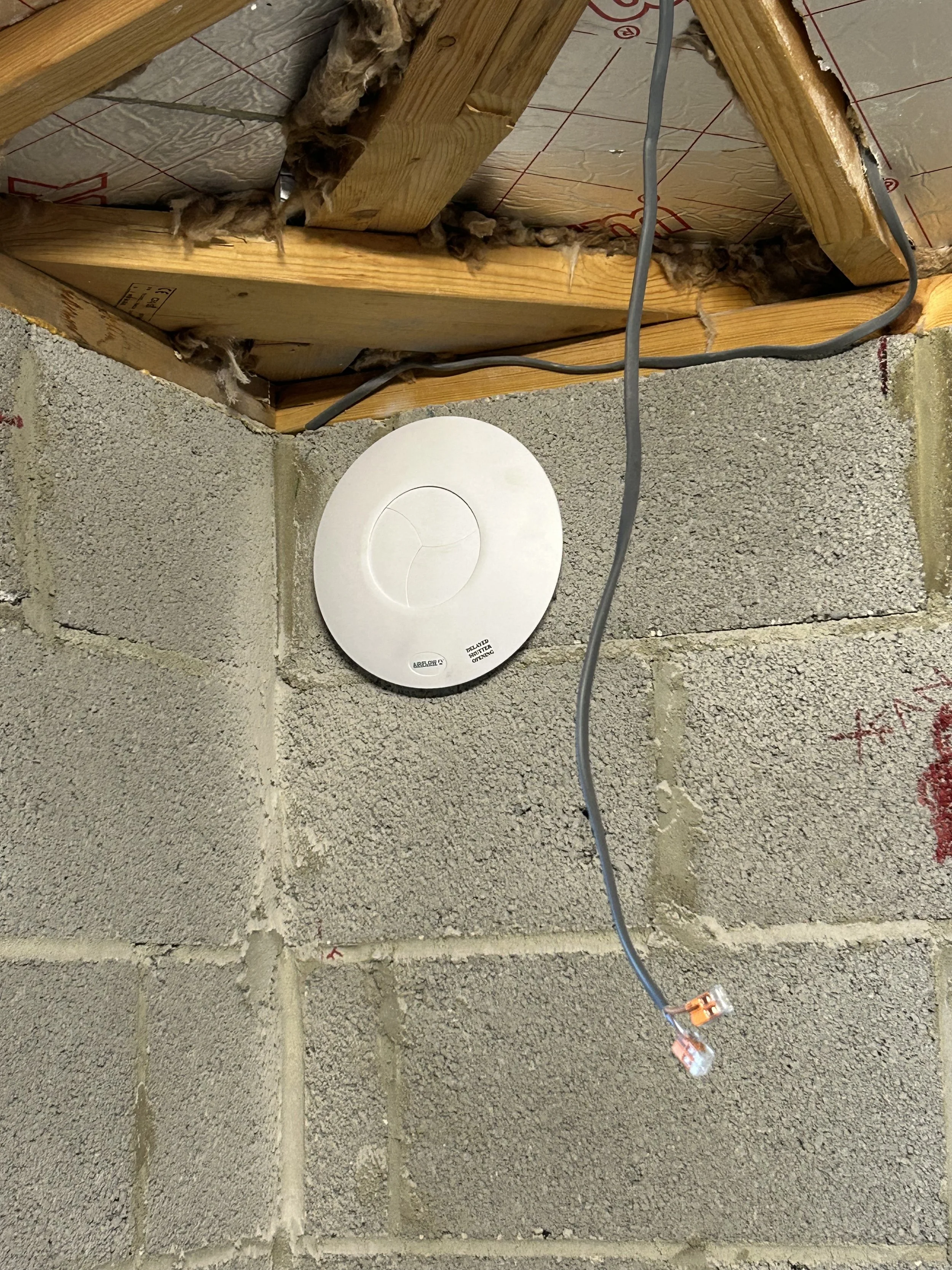 A smoke detector mounted on a cinder block wall in a basement ceiling with exposed wooden framing and electrical wires.