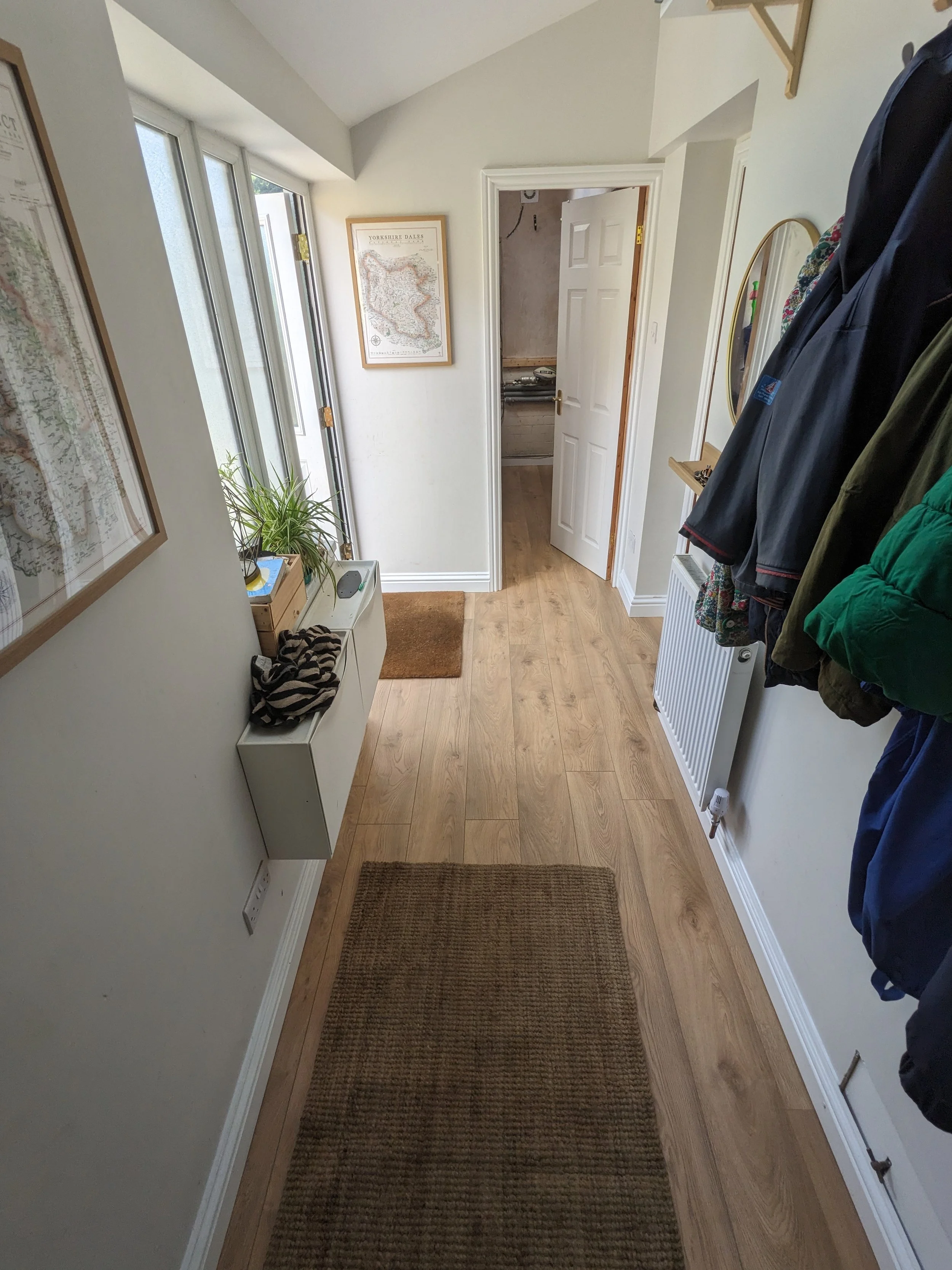 View of an entryway with wooden flooring, a brown rug, a radiator on the right wall, coats hanging, a small white shelf on the left wall, and a framed map of Yorkshire Dales on the wall. An open door leads to another room.