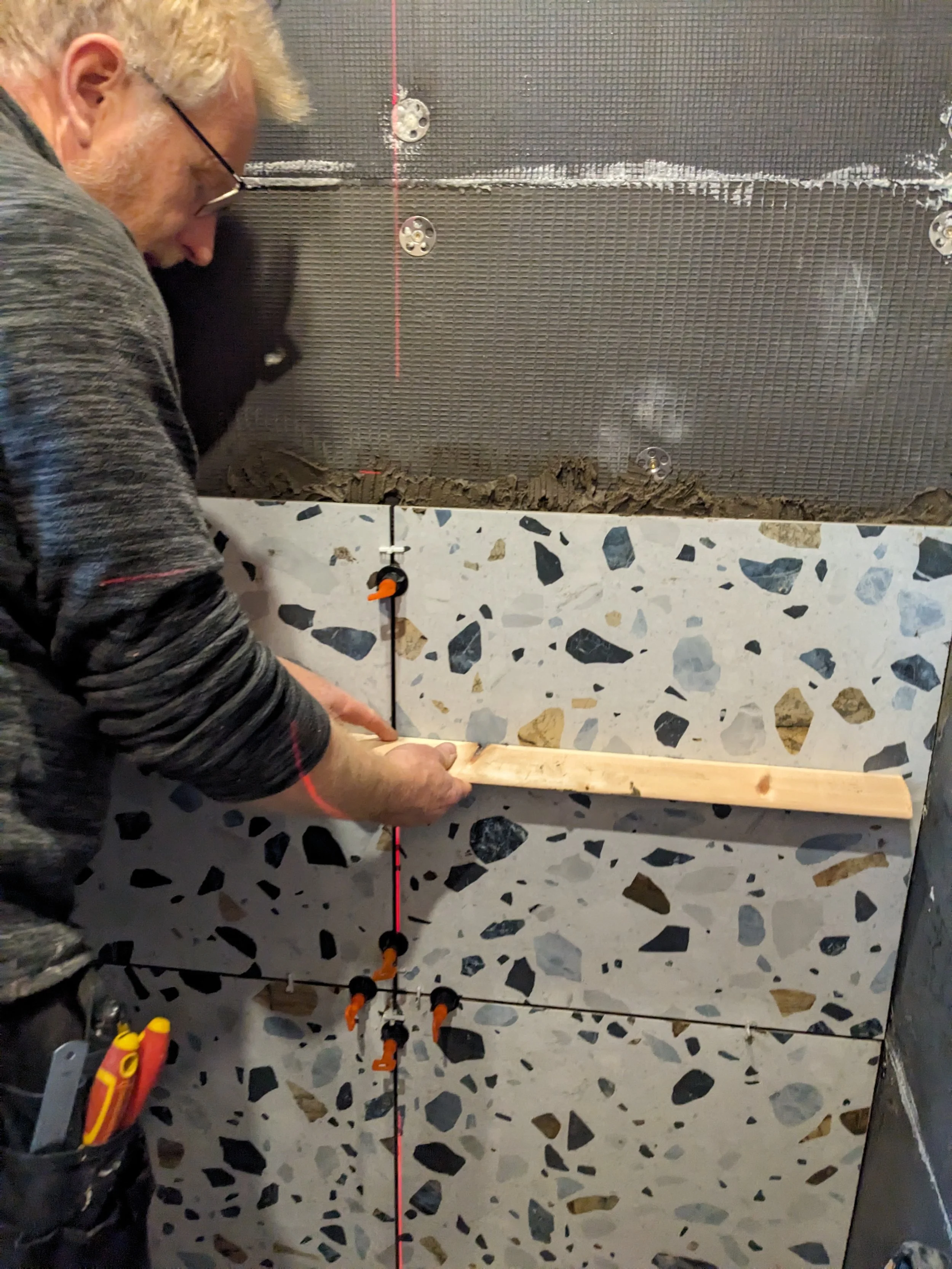 A man working on tiling a wall, using a level to ensure tiles are straight, with tile spacers between the tiles.