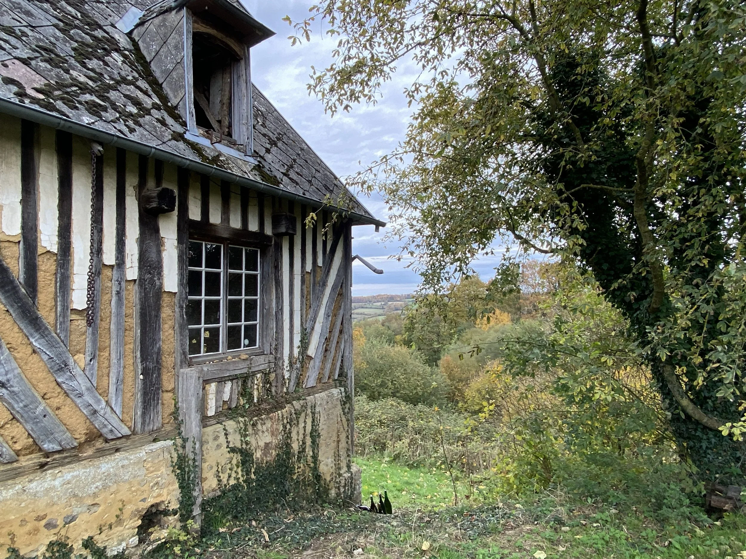 Une maison ancienne en pierre et bois avec un toit en ardoise, entourée d'arbres en automne, dans un paysage rural