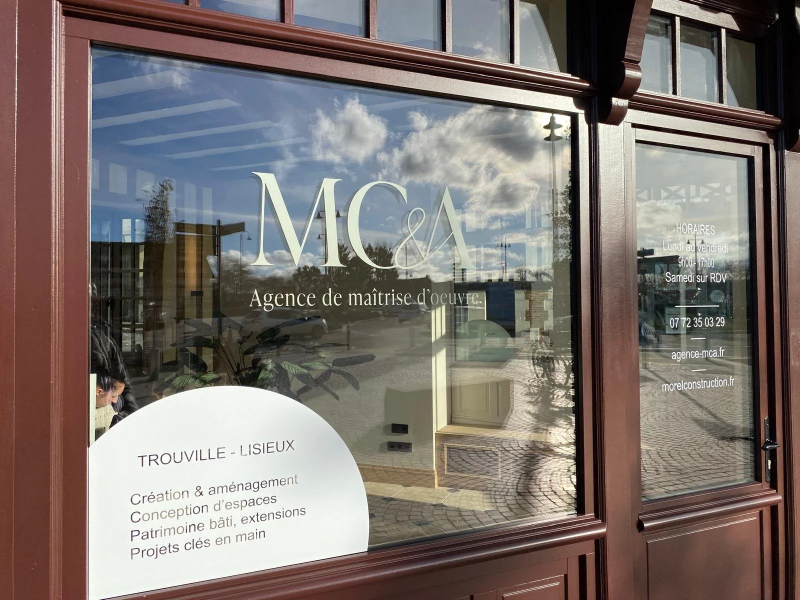 Vitrine d'une agence de maîtrise d'œuvre nommée MCA, située à Trouville-Lisieux, avec horaires, contact et site web, reflet du ciel avec quelques nuages et une rue piétonne à l'extérieur.
