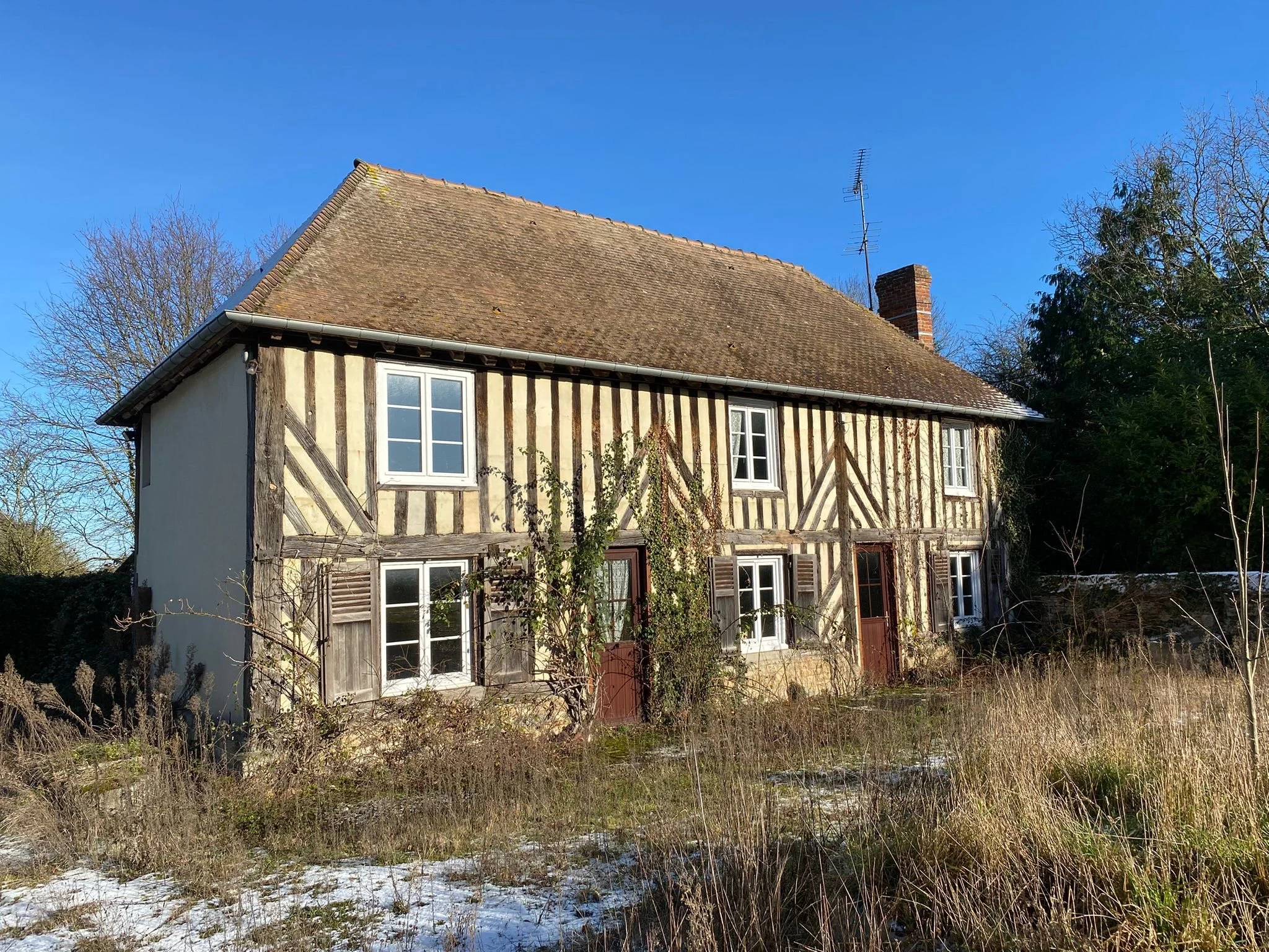 Maison ancienne à colombages avec un toit en tuiles, située dans un environnement rural. Le terrain environnant est en friche, avec de l'herbe sèche et quelques branches d'arbres sans feuilles.
