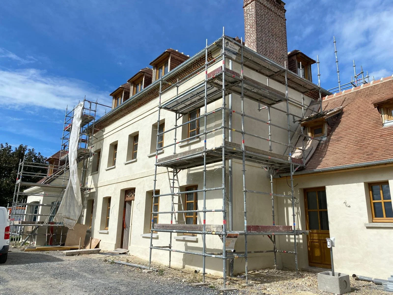 Chantier de construction d'une maison avec échafaudages en métal et façade en cours de rénovation, fenêtres en bois, sous un ciel bleu avec quelques nuages.