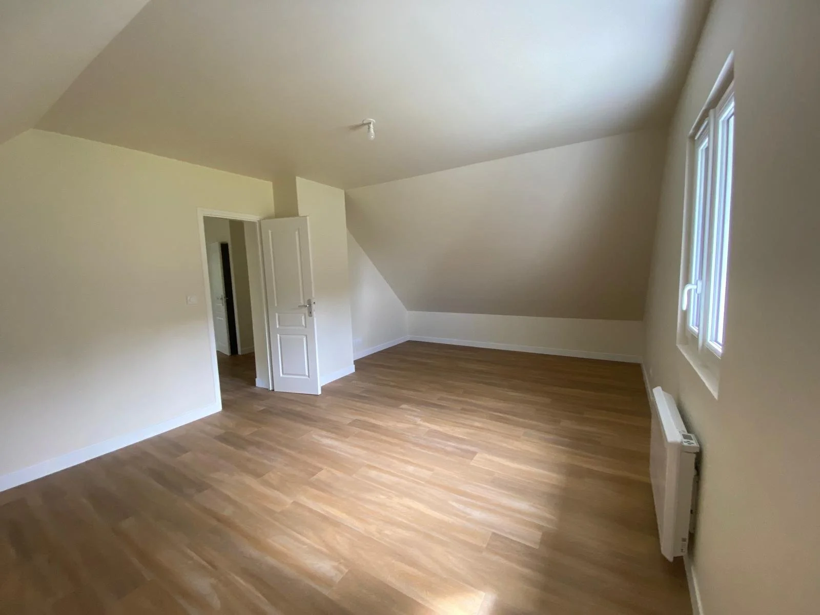 Pièce vide avec des murs crème, deux fenêtres, un radiateur, un sol en bois clair et une porte blanche, plafond incliné.