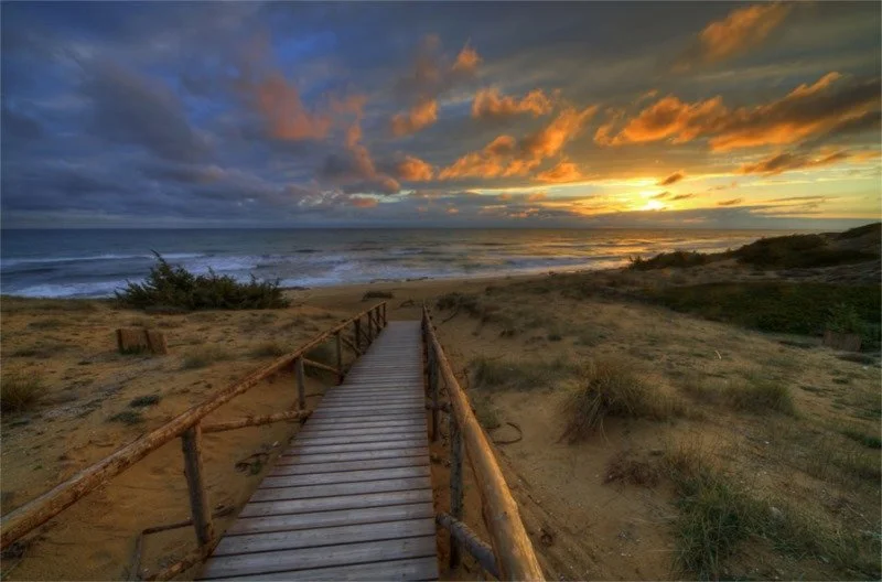 Wooden pathway leading to a beach with sand dunes, grasses, and ocean under a colorful sunset sky with clouds.