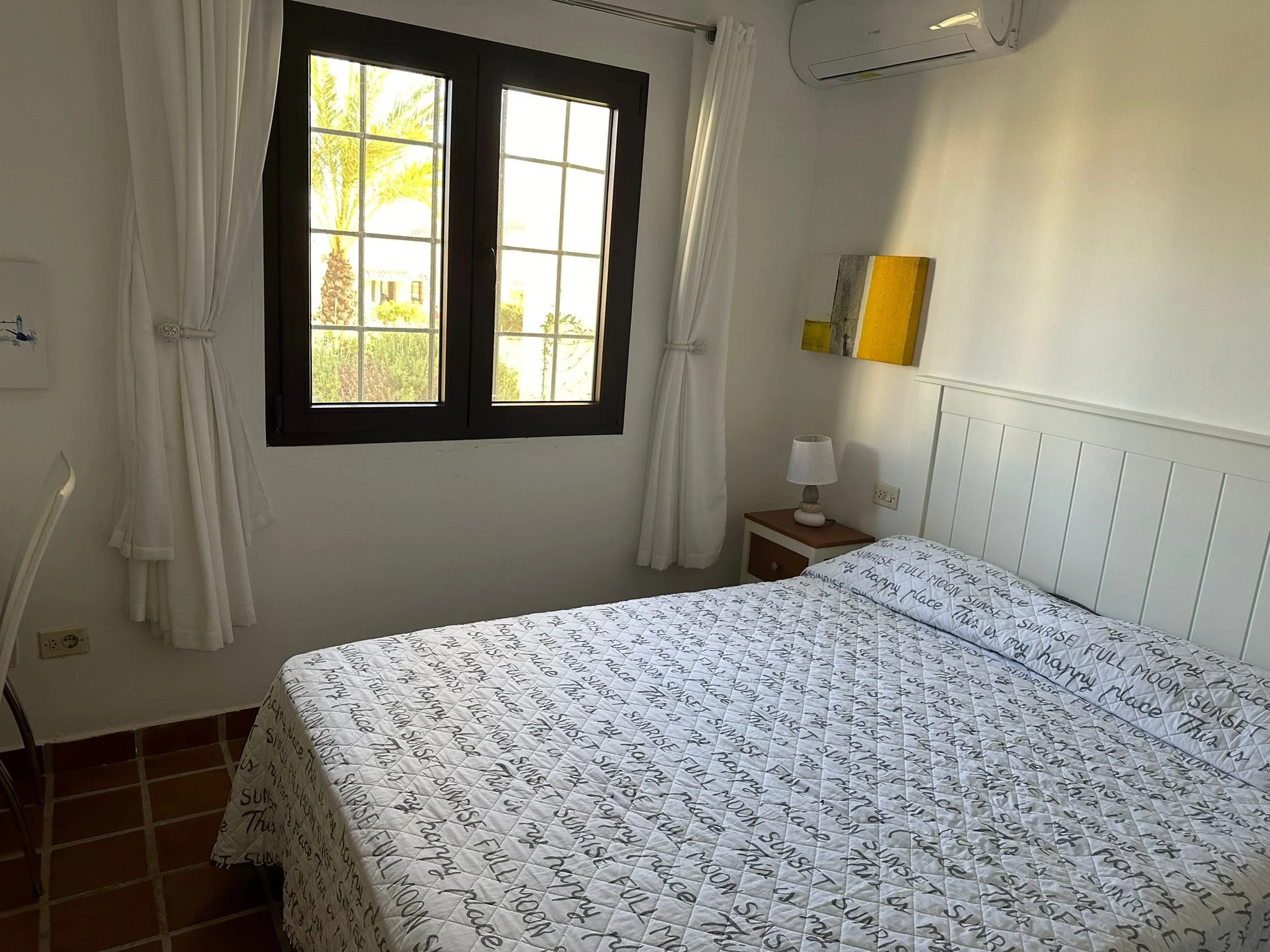 A bedroom with a bed covered in a white quilt with black handwritten text, a nightstand with a table lamp, a window with white curtains, a small abstract wall art, and an air conditioning unit mounted on the wall.