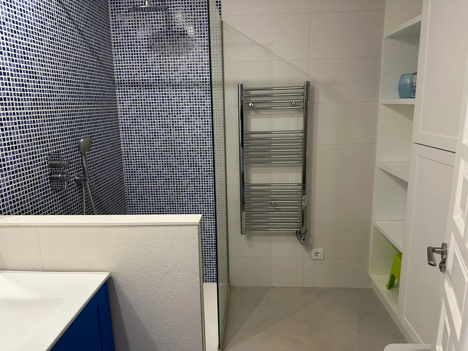 Bathroom with a walk-in shower featuring navy blue mosaic tiles, a handheld showerhead, a white shelf with a blue container, a chrome heated towel rail, a white cabinet, and a light-colored floor.