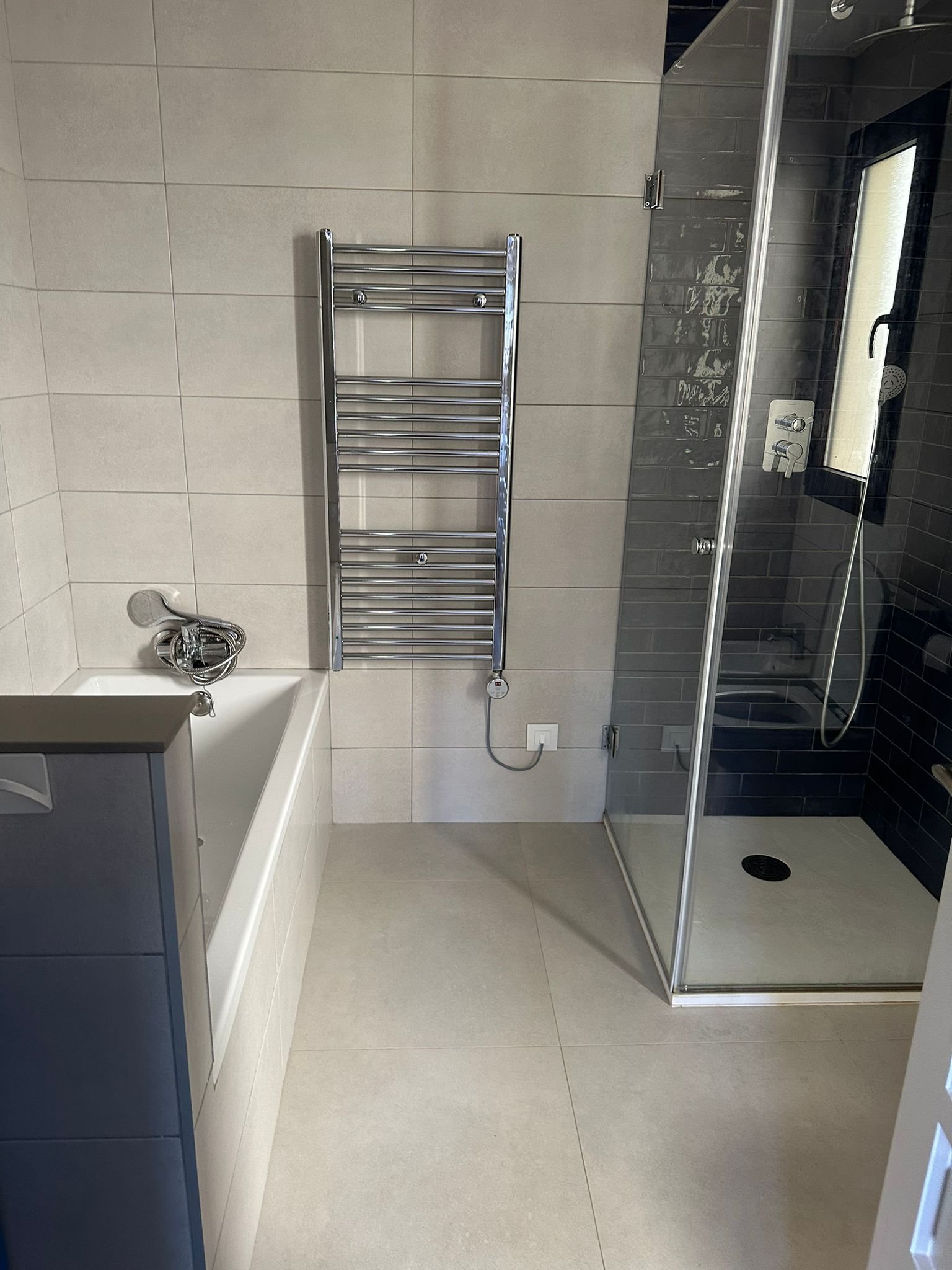 Modern bathroom with beige tiled floor and walls, a white bathtub with a handheld showerhead, a chrome towel rack, a shower with a glass enclosure, and dark gray tiling inside the shower area.