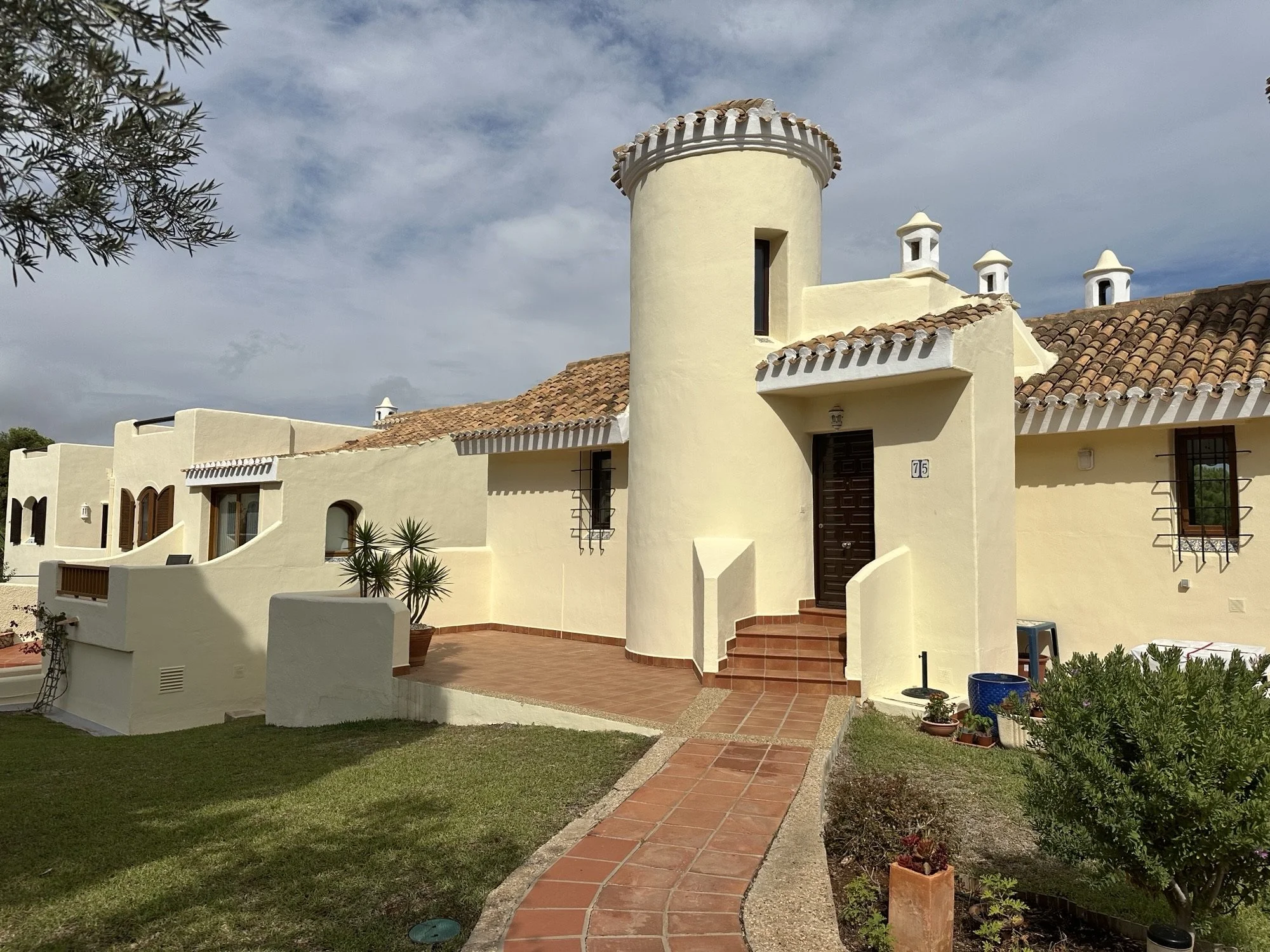 A row of Mediterranean-style houses with yellow stucco walls, terracotta roof tiles, and decorative chimney stacks. A pathway leads to one house entrance, and there are small potted plants and a garden area in the yard.