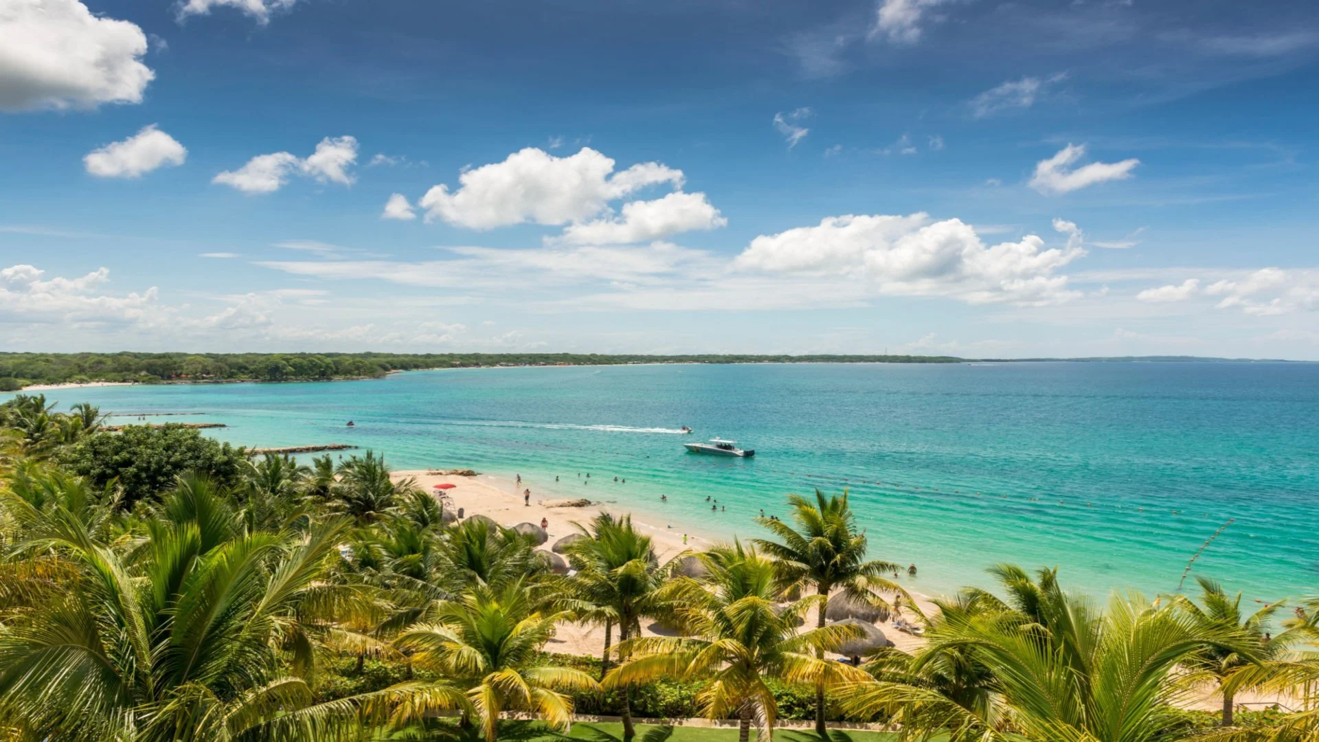 Tropical beach with white sand, palm trees, turquoise water, and boats in the distance, under a partly cloudy sky.