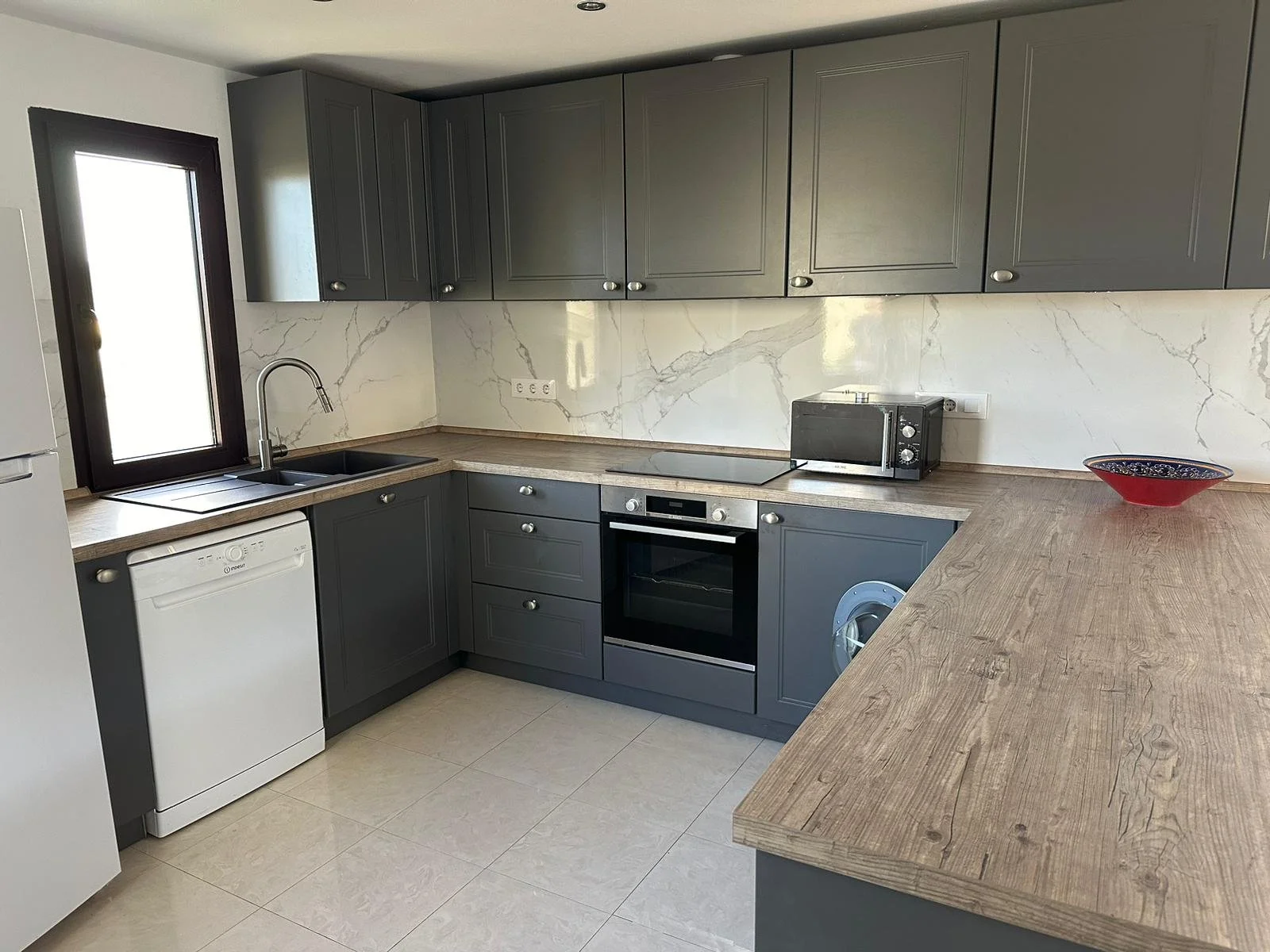 Modern kitchen with gray cabinets, beige marble backsplash, black sink, oven, small microwave, dishwasher, and a wooden countertop with a red dish and a toaster.