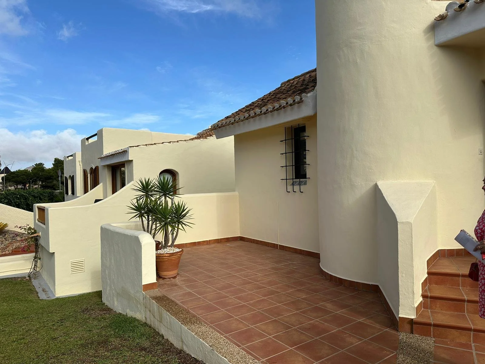 White Mediterranean-style house with curved walls, terracotta tiled patio, potted plant, and a person holding a tablet on the right side.