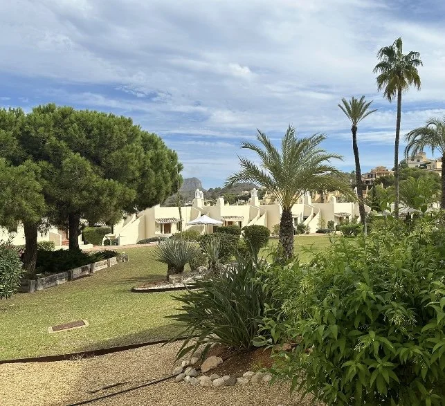 Lush green park with various trees and plants, white buildings in the background, and a partly cloudy sky overhead.