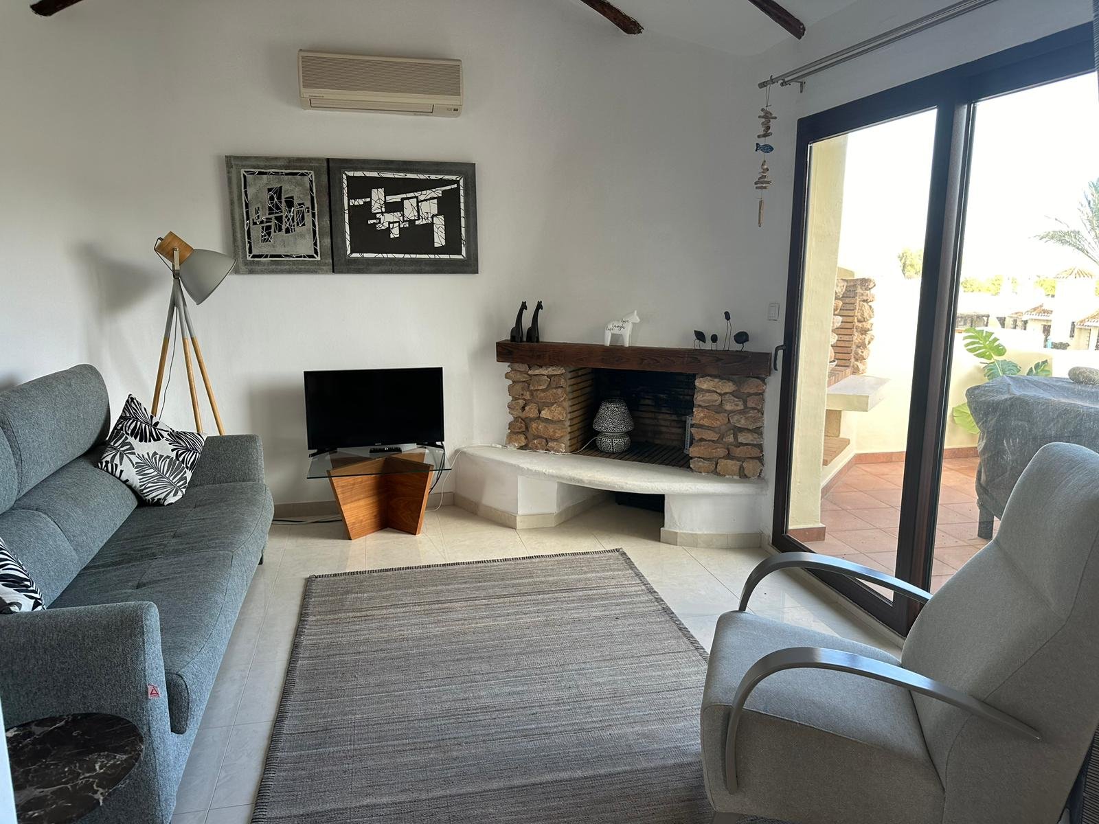 Living room with gray sofa and patterned pillows, a floor lamp, a glass TV stand with TV, a brick and wood fireplace, sliding glass door leading to a balcony, and wall decorations.