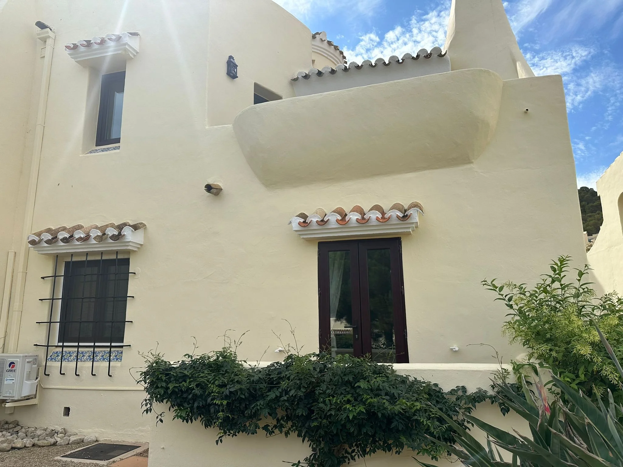 A white Mediterranean-style house with curved walls, tiled roof accents, and barred windows, surrounded by greenery and set against a blue sky with some clouds.