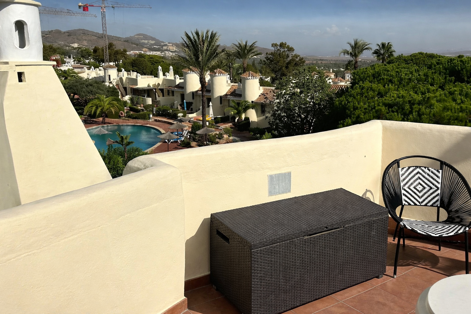 Balcony view overlooking a resort with a swimming pool, palm trees, white buildings with terracotta roofs, and mountains in the background.