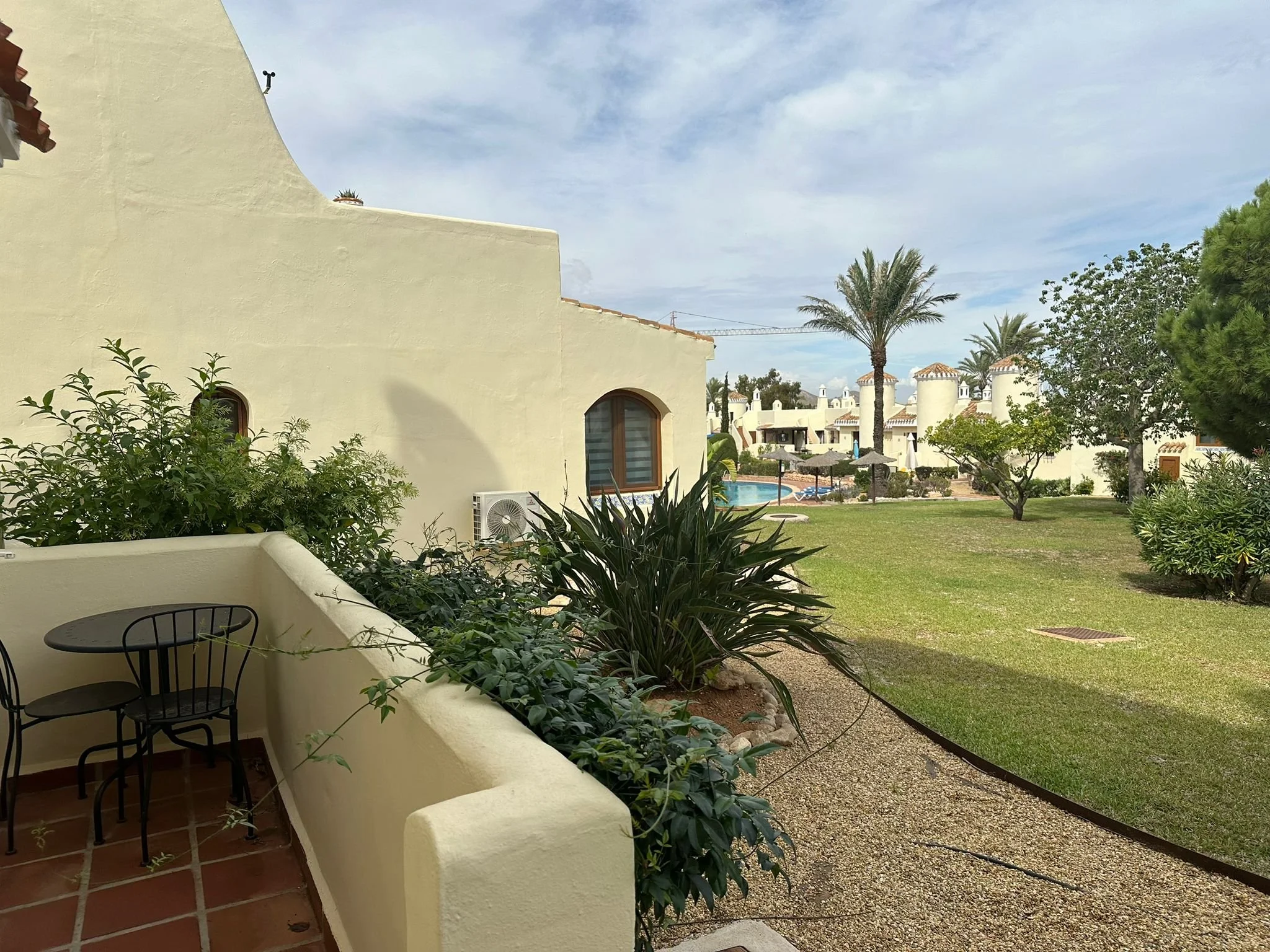 Balcony with two black chairs and a small table, surrounded by green plants and overlooking a green lawn with trees and a pool area with umbrellas, palm trees, and white buildings in the background.