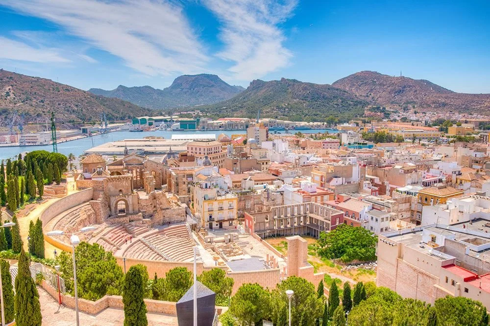 A cityscape with ancient ruins, colorful buildings, a body of water, and mountains in the background under a partly cloudy sky.