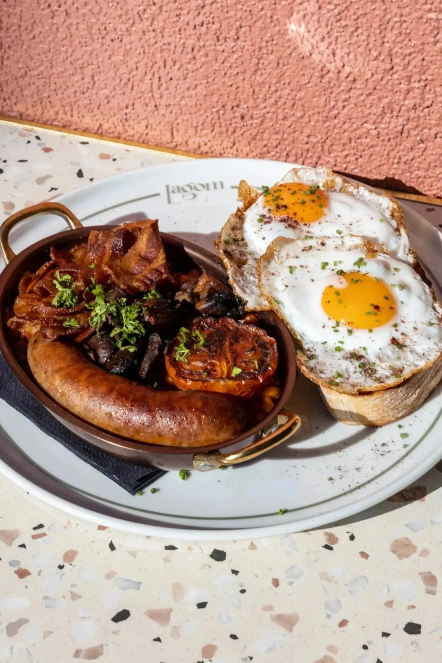 A plate of breakfast food including two fried eggs, fried bacon, and sausages, placed on a table with a pink carpet in the background.